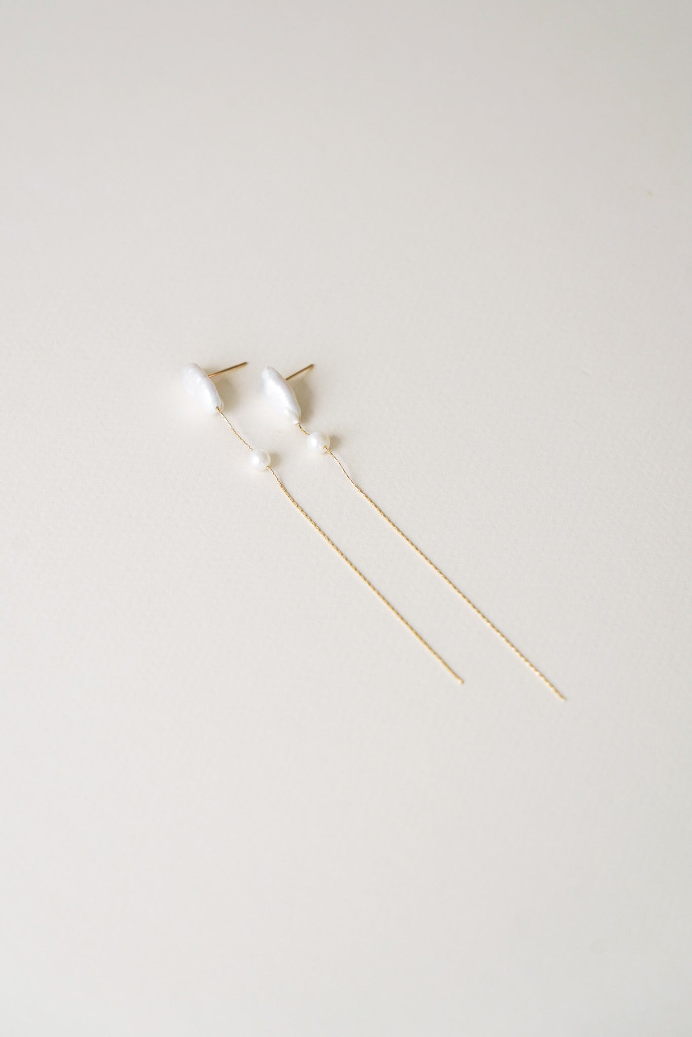 Paire de boucles d’oreilles longues photographiée sur fond clair, composée d’une perle de culture blanche de taille généreuse, suivie d’une perle plus petite en délicat contraste. Le design fin et épuré, aux lignes élancées, évoque la légèreté et l’élégance naturelle. Une création subtilement sophistiquée, idéale pour apporter une touche de grâce discrète à une tenue de mariage ou de cérémonie.