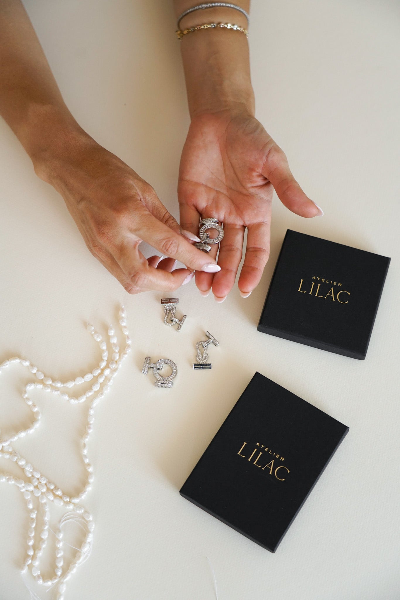 Une main habile est en train de fabriquer un bijou, tenant délicatement une partie du bijou. On peut distinguer plusieurs perles colorées ainsi que différentes attaches métalliques utilisées pour l’assemblage du bijou. En arrière-plan, plusieurs boîtes portant le logo de l’atelier Lilac, montrant l’endroit où les bijoux finis sont soigneusement emballés et prêts à être expédiés.