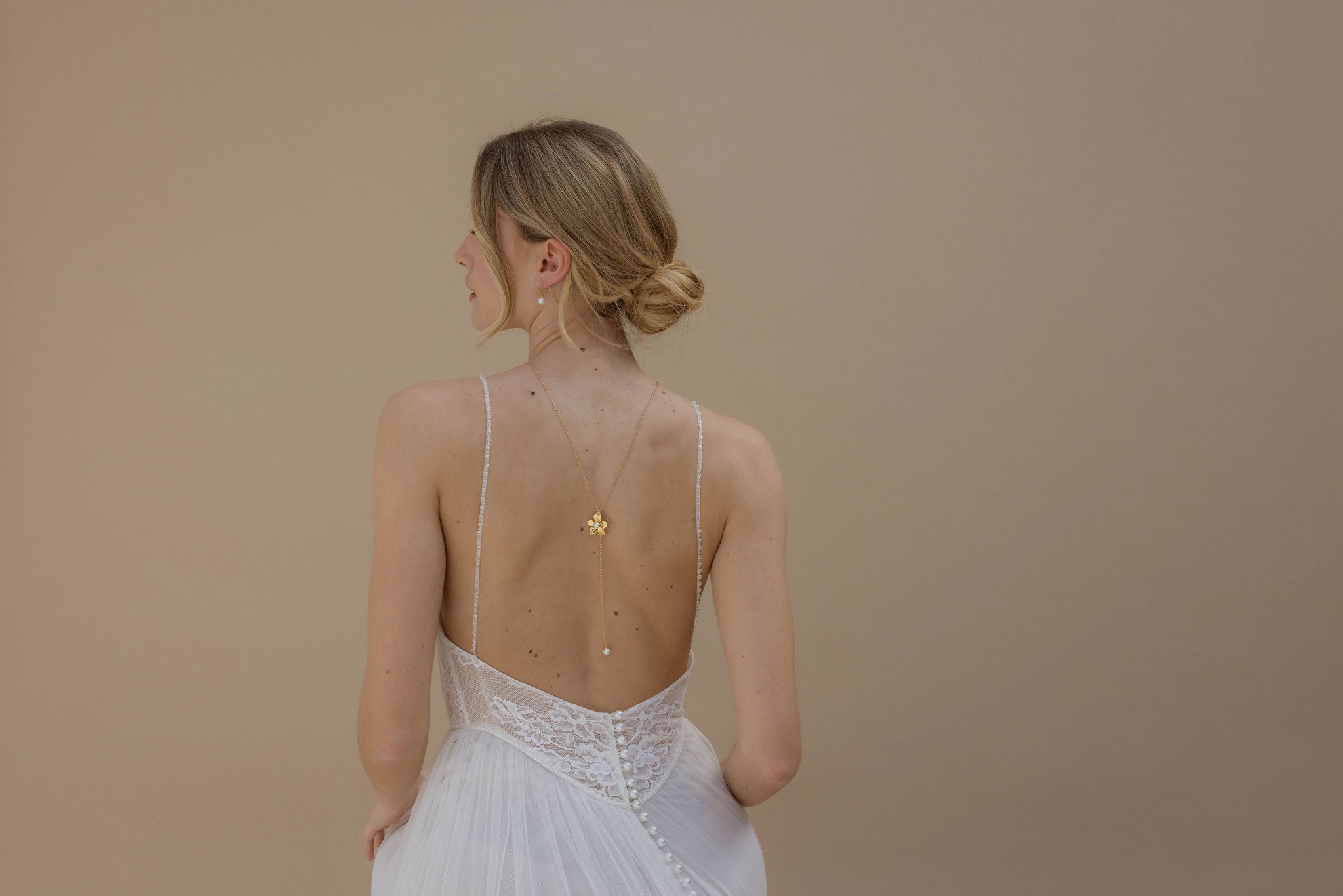 Mariée vue de dos, portant une robe en dentelle à fines bretelles avec une fermeture perlée. Elle a un joli collier orné d’une fleur et d’une perle tombant le long de son dos. Sa tête est tournée sur le côté, avec un chignon bas et des mèches délicates encadrant son visage. Elle porte des boucles d’oreilles courtes ornées de perles.
