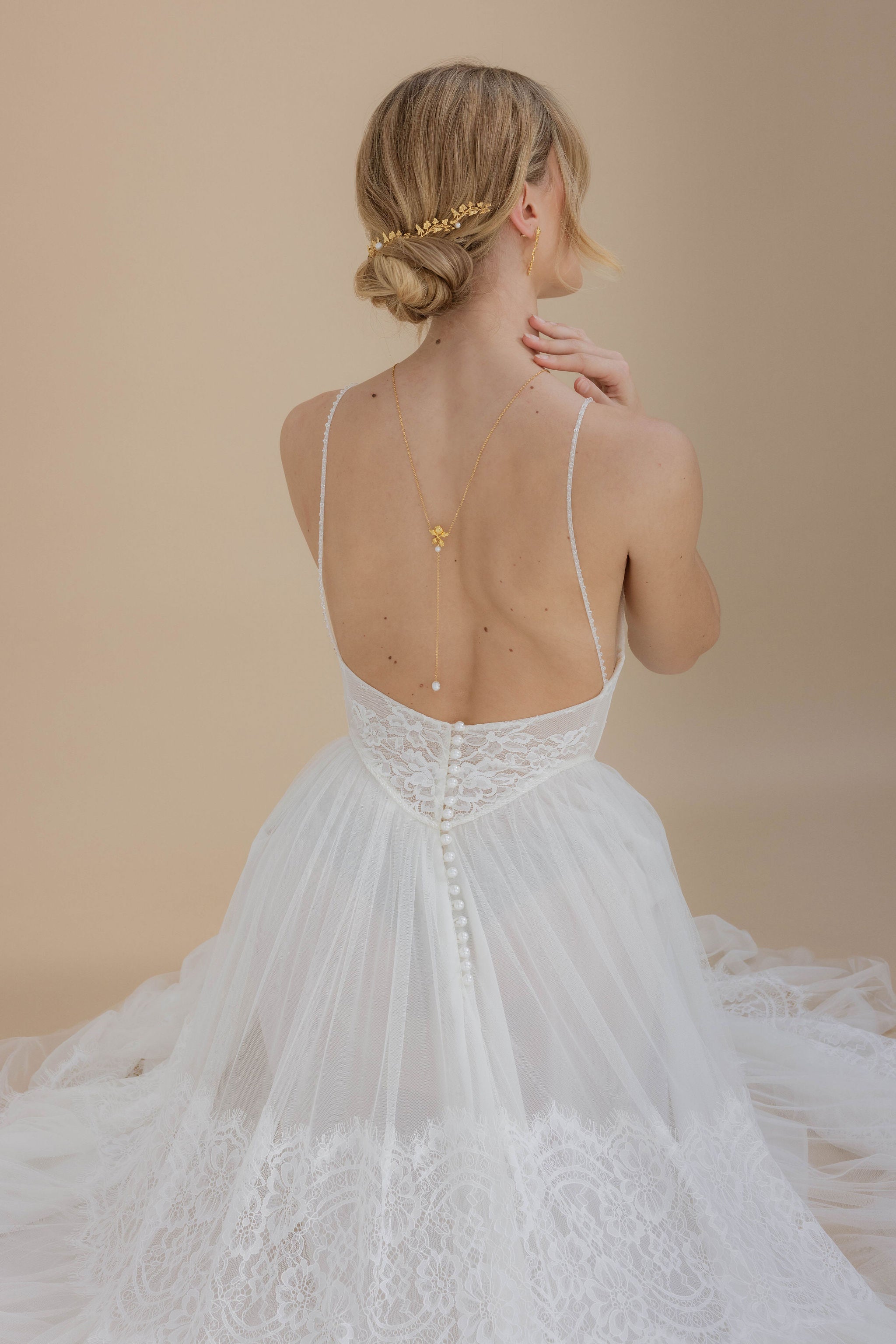 Mariée au style romantique coiffée d’un chignon bas, sublimé par une vigne dorée, avec un collier de dos raffiné et des boucles d’oreilles florales pour un ensemble harmonieux.