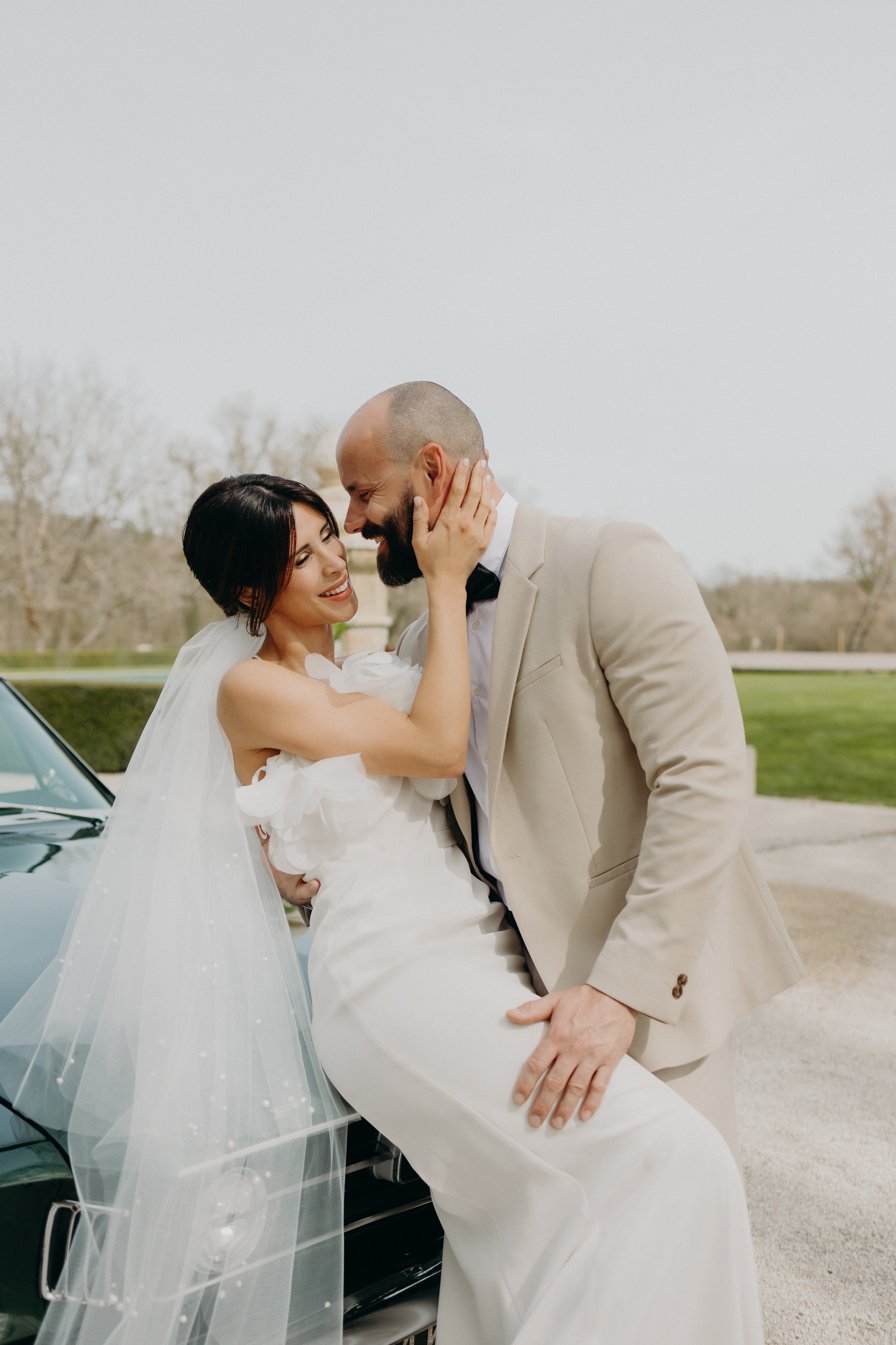 Un couple de jeunes mariés modernes et élégants s’enlace tendrement devant une voiture vintage, sous une lumière douce, lors de leur réception de mariage au décor aux nuances matcha et olive, mêlant élégance naturelle et romantisme délicat. Inspirations mariage naturel et moderne sue le thème matcha olive.