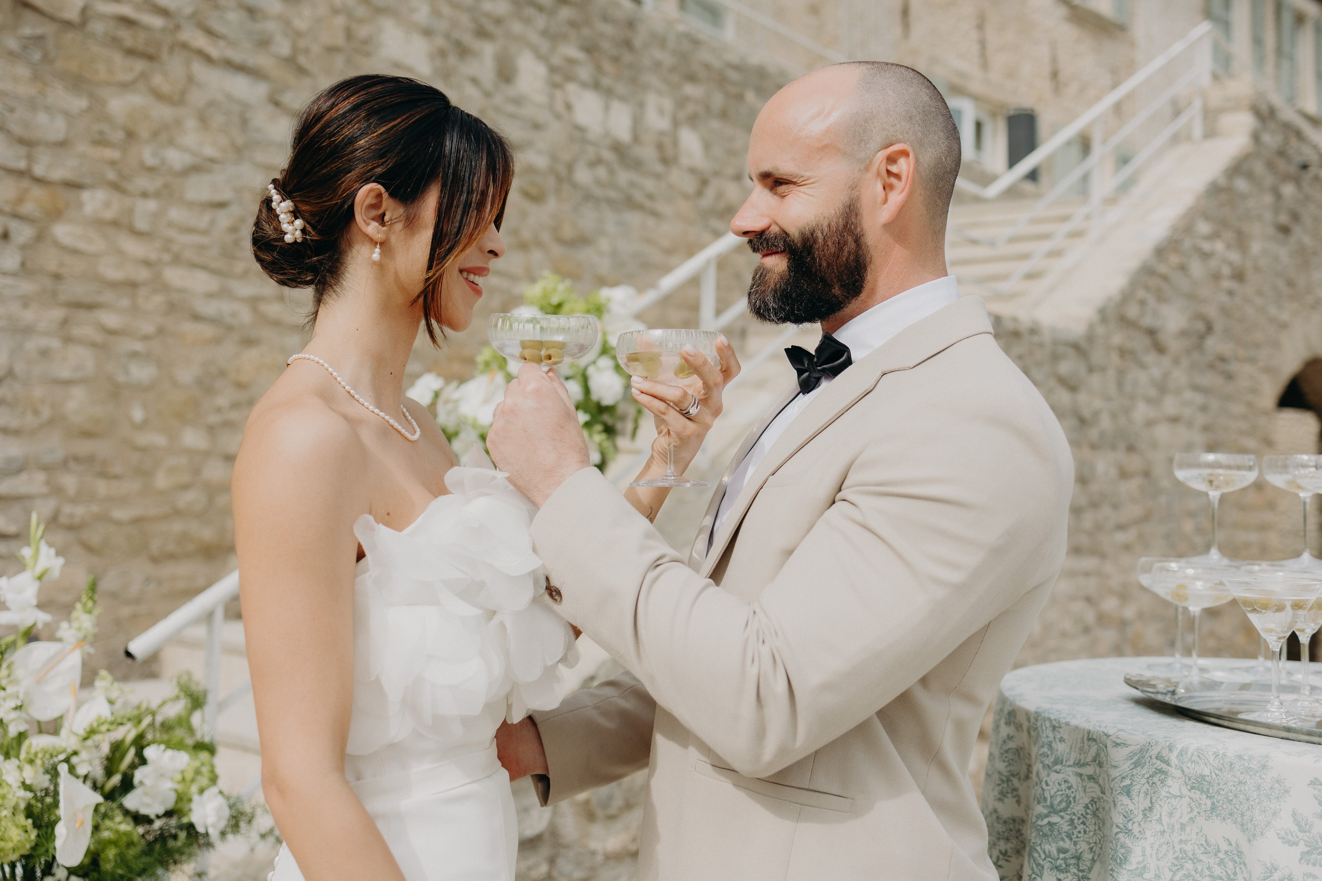 Couple élégant qui se donnent à boire dans une belle coupe lors de leur mariage naturel aux inspirations matcha et olive, on peut voir une tour de coupes de champagne à droite et des fleurs blanches à gauche, la réception est moderne dans un chateau aux pierres naturelles avec de la verdure qui rappelle le thème matcha et olive.
