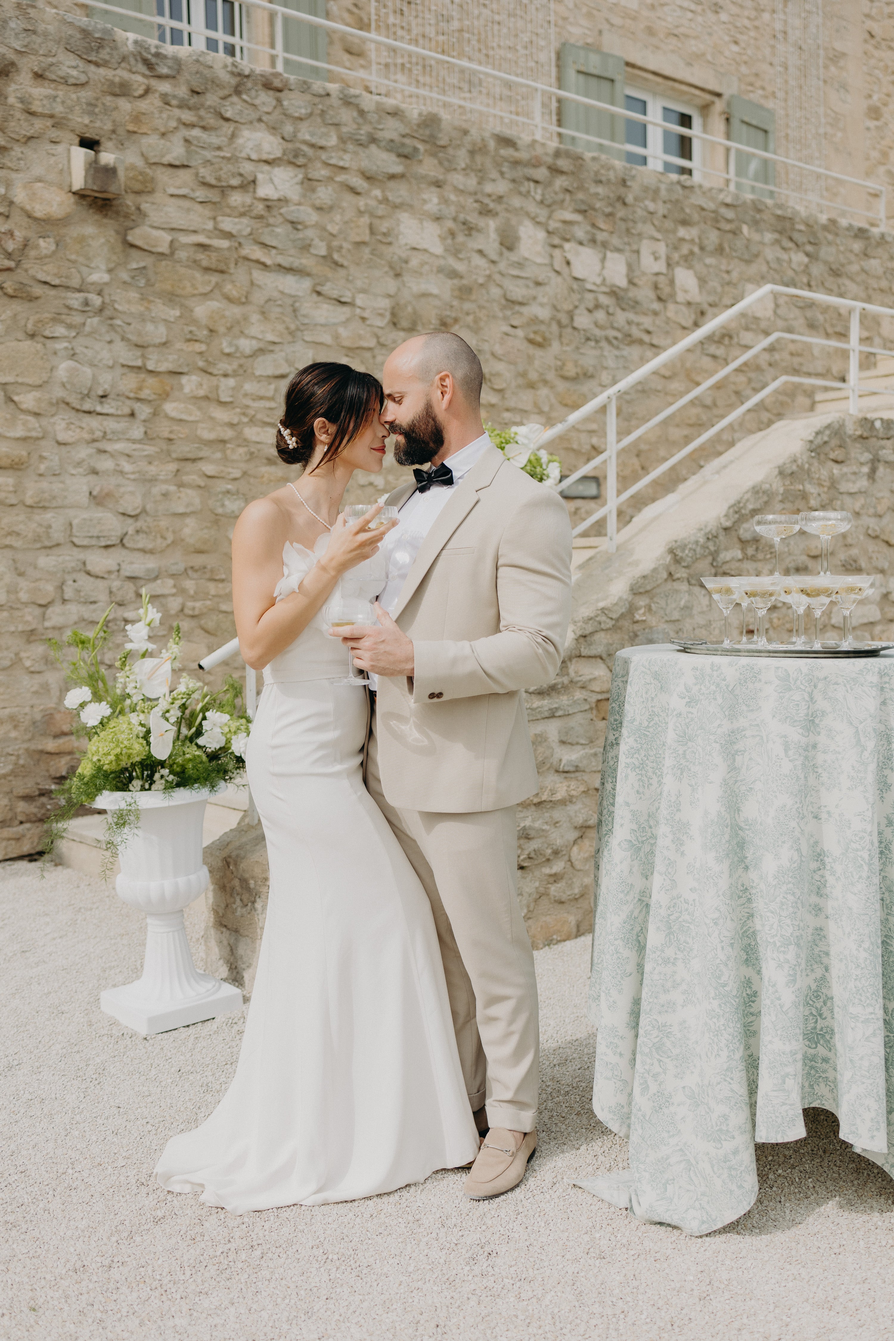 Couple élégant qui s'enlace lors de leur mariage naturel aux inspirations matcha et olive, on peut voir une tour de coupes de champagne à droite et des fleurs blanches à gauche, la réception est moderne dans un chateau aux pierres naturelles avec de la verdure qui rappelle le thème matcha et olive.