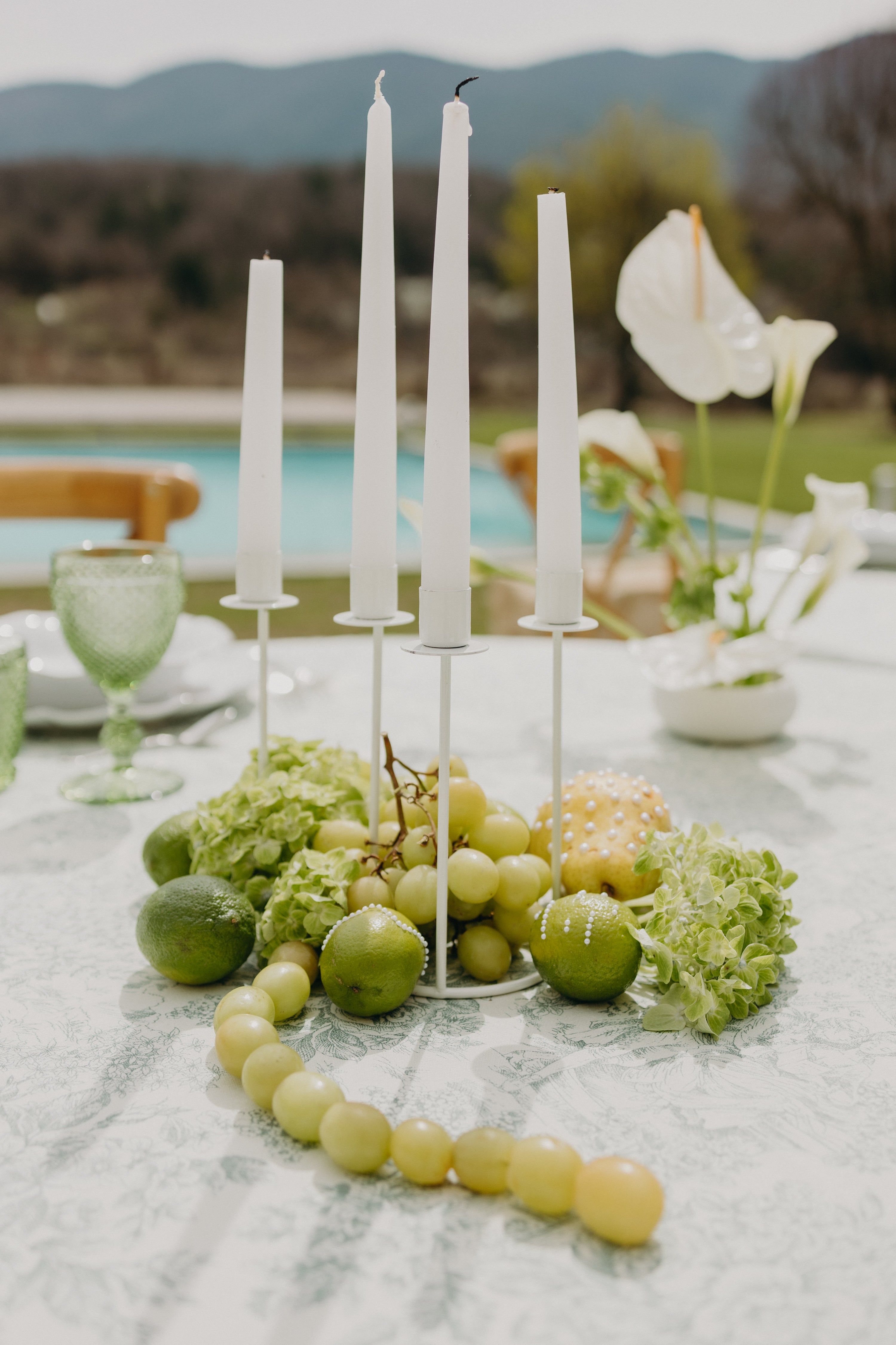 Décoration de table moderne et vives aux couleurs vertes rappelant le thème matcha et olive, bougies blanches et fruits verts accompagnés de verdure au centre de la table, on peut voir des fleurs, des chaises et une piscine dans ce château idéal pour un mariage inspiration matcha et olive, moderne et naturel