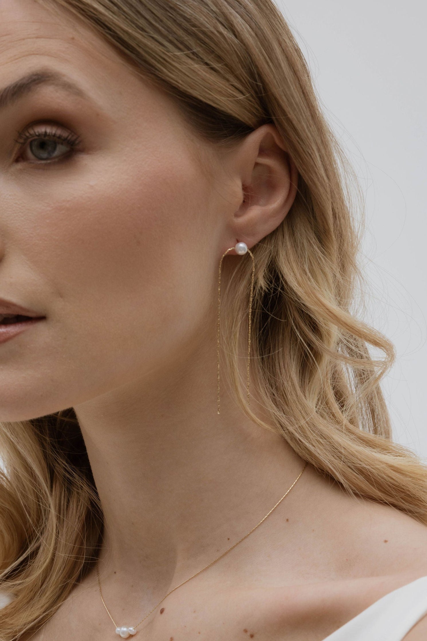 Focus sur l'oreille d'une mariée portant des boucles d'oreilles pendantes avec une chaine et une perle naturelle de culture minimaliste