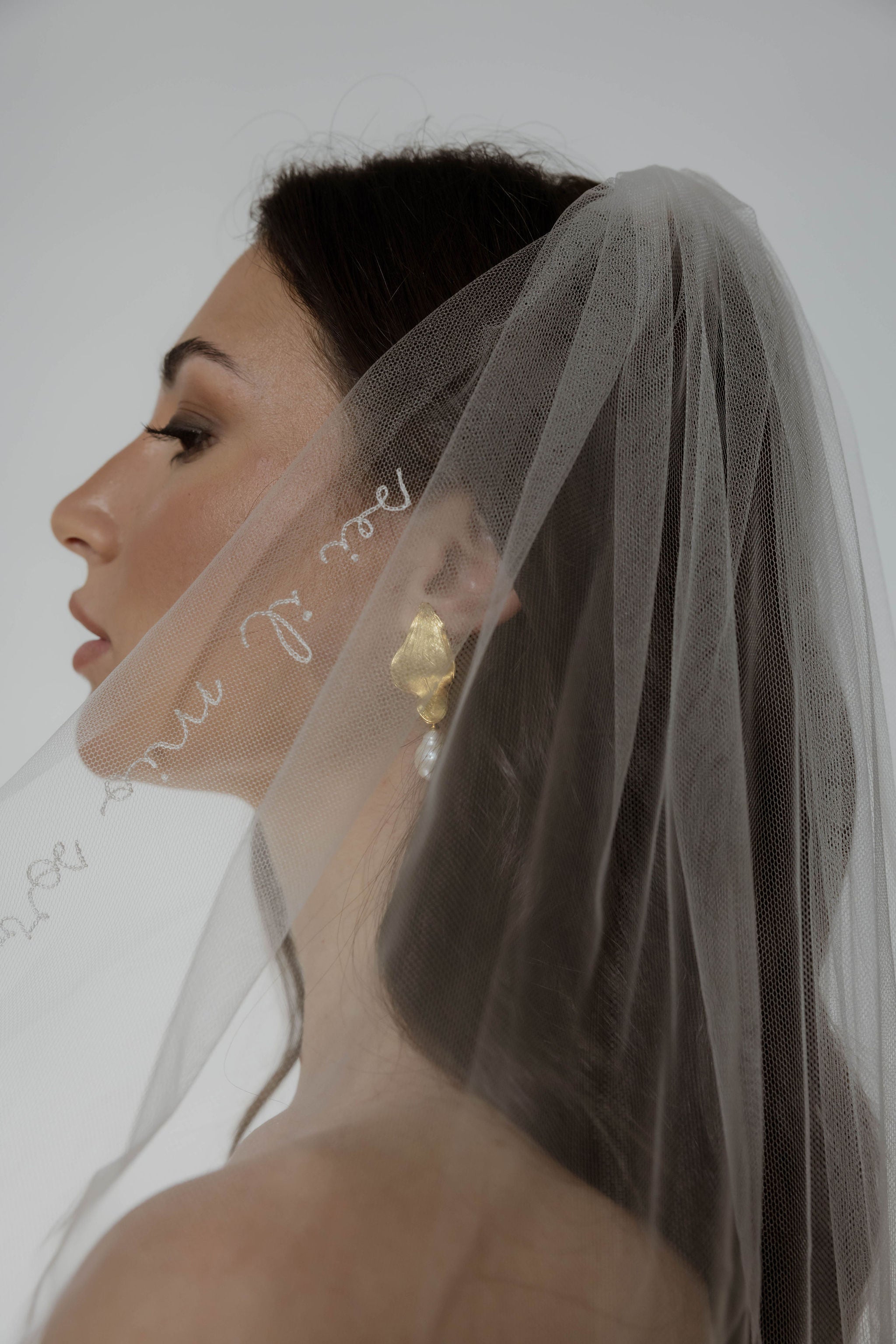 Mariée de profil portant un voile brodée et derrière on peut voir des boucles d'oreilles pendantes en forme de feuille