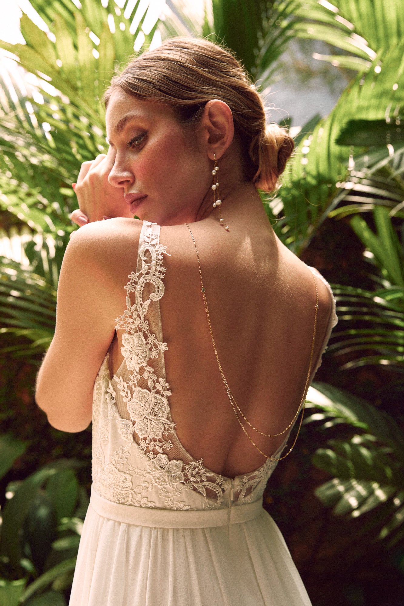 Mariée dans un jardin tropical coiffée d'un chignon bas bohème, avec des boucles d'oreilles pendantes élégantes à perles et collier de dos bohème à doubles chaînes et cristaux