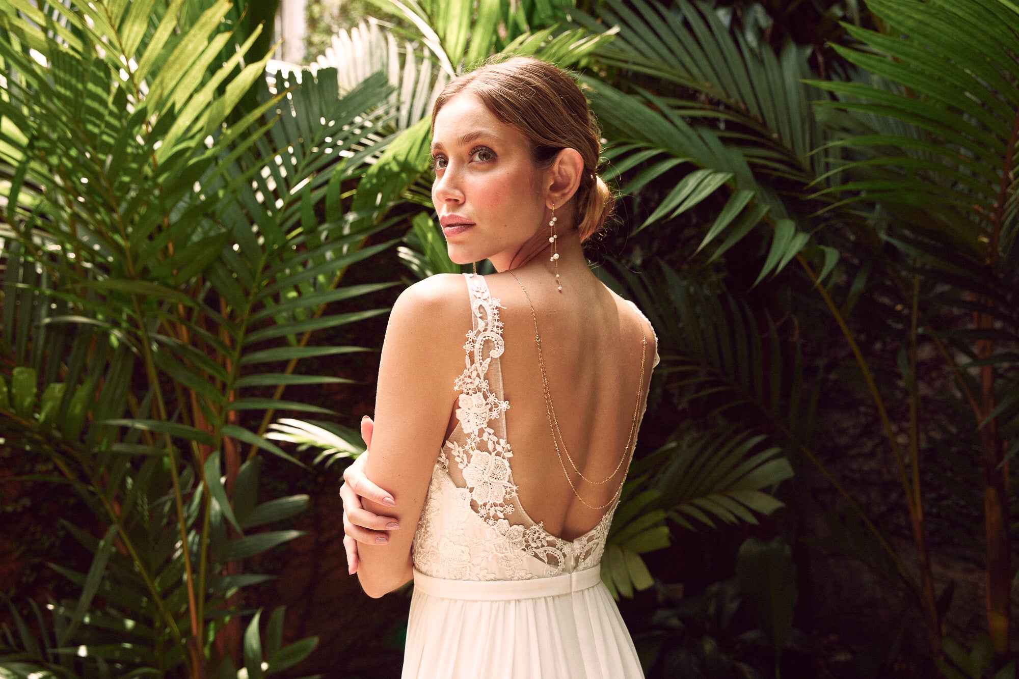 Mariée avec un chignon bohème portant un collier de dos à double chaine et des boucles d'oreilles pendantes à perles devant un décor tropical