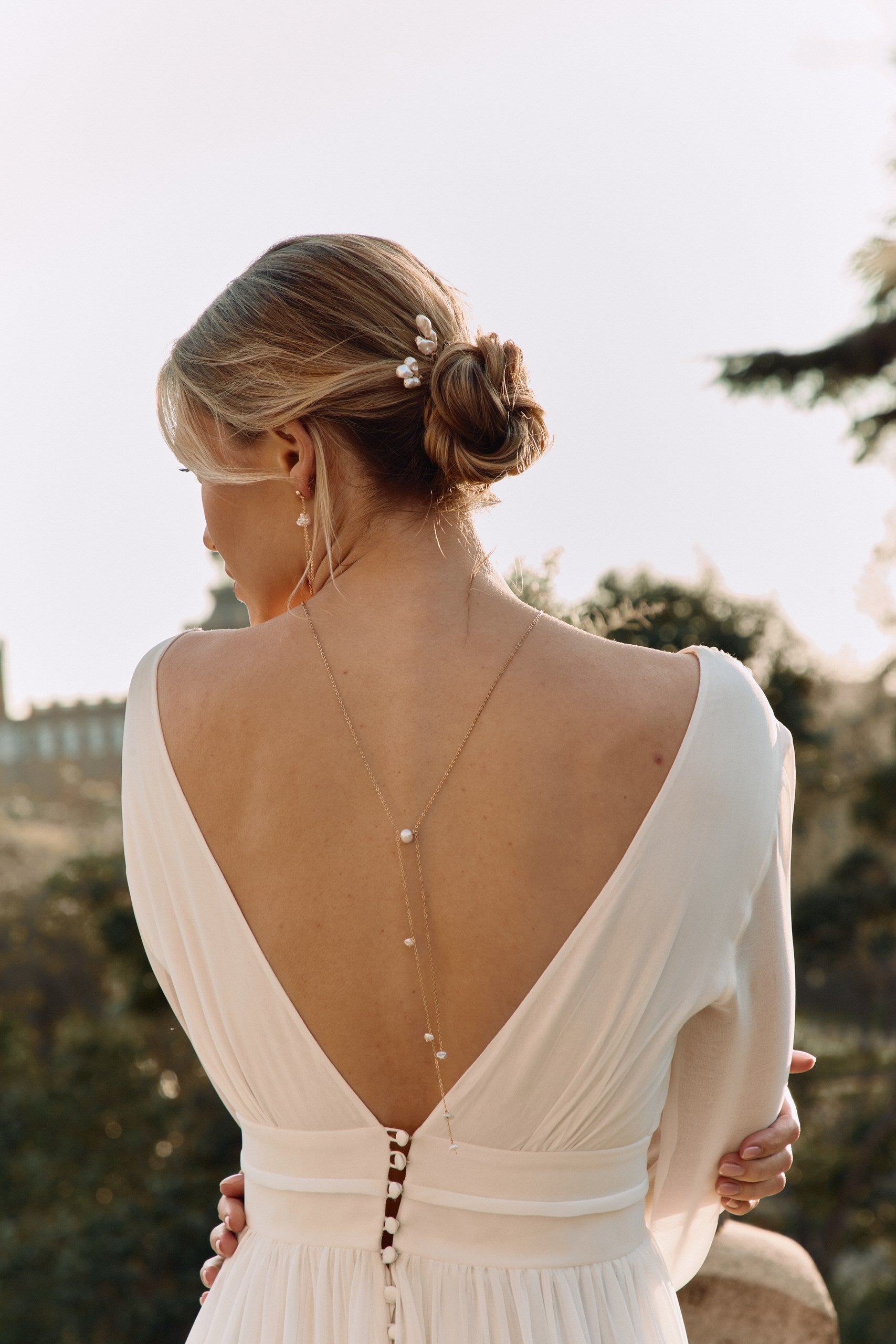 Mariée de dos portant une robe bohème qui à un grand décolleté de dos avec un collier de dos fait de perles naturelles et des piques dans les cheveux 