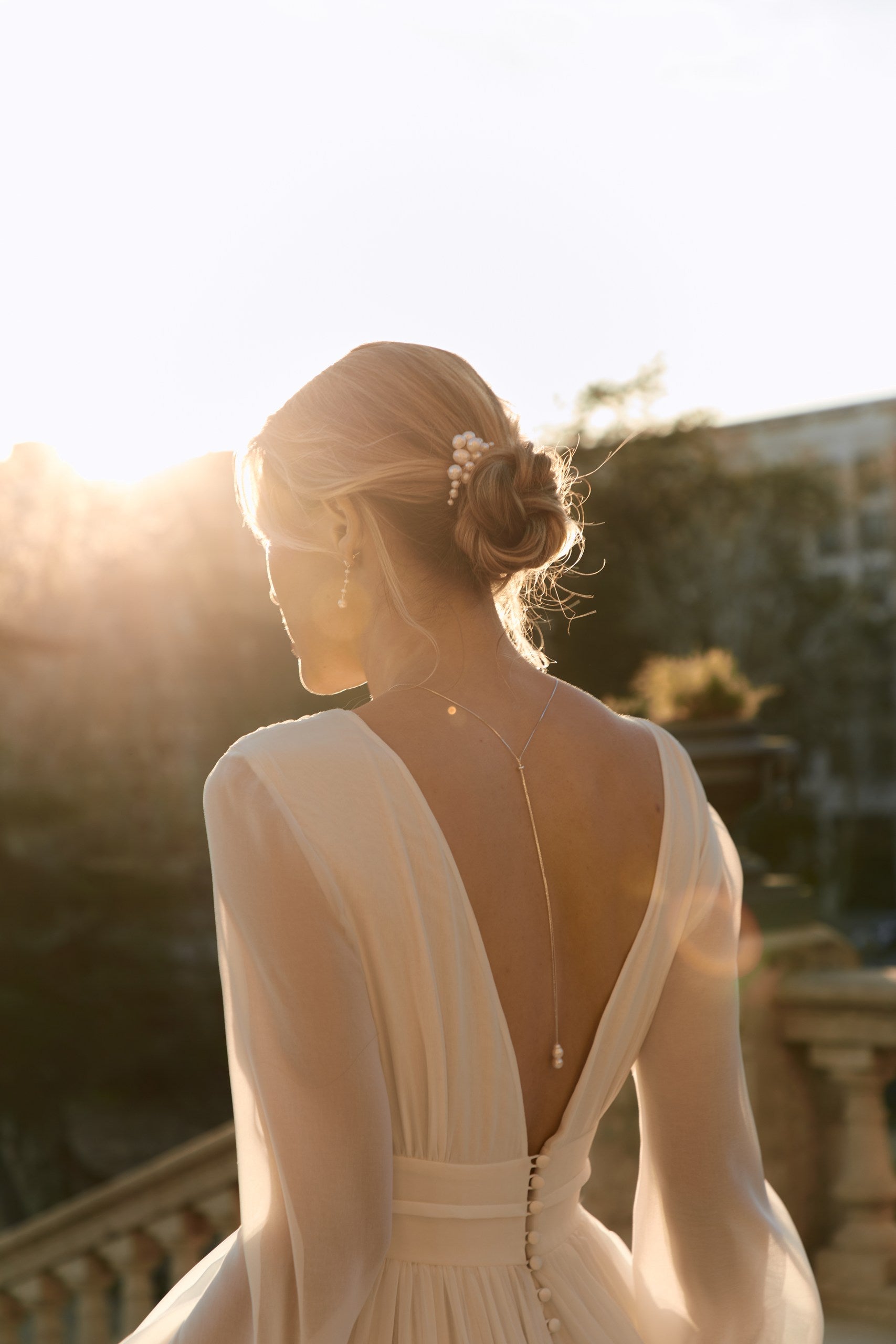 femme de dos portant une robe de mariée à manches longues avec un décolleté de dos et des bijoux tel qu'un collier de dos, une paire de boucle d'oreilles et une peigne en perles