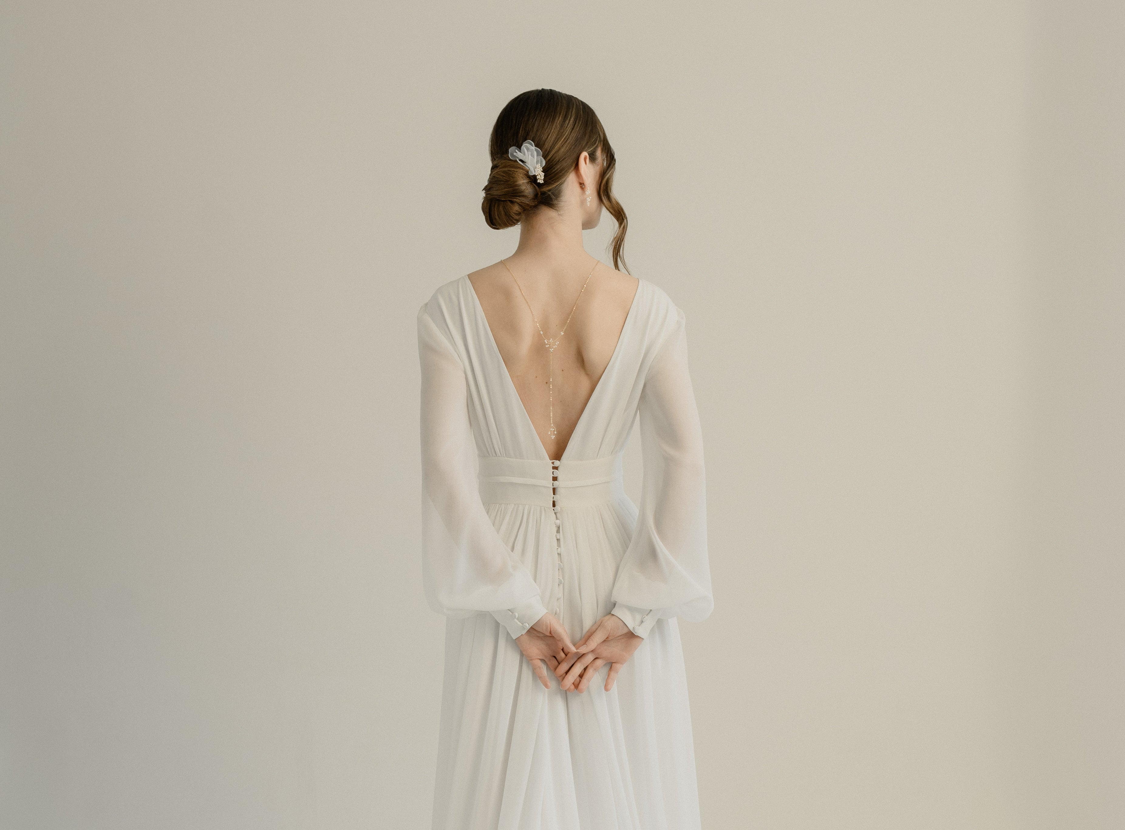 Jeune femme de dos avec les mains qui se touchent dans une robe de mariée coiffé avec un chignon montrant un collier de dos délicat et des accessoires de cheveux 