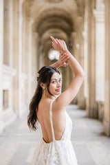 Mariée sous les arches de la place du Louvre à Paris portant des boucles d'oreilles pendant avec une fleur et des chaines en or et un pic à cheveux assorti