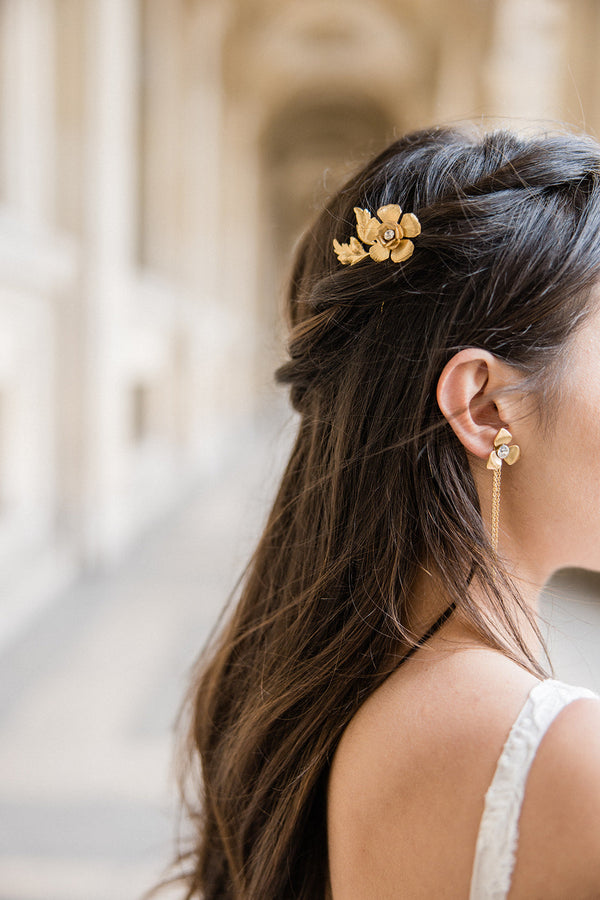 Mariée avec une demi-queue tressée au style bohème coiffée d'un pic à cheveux à fleur doré et portant des boucles d'oreilles pendantes dorées assorties