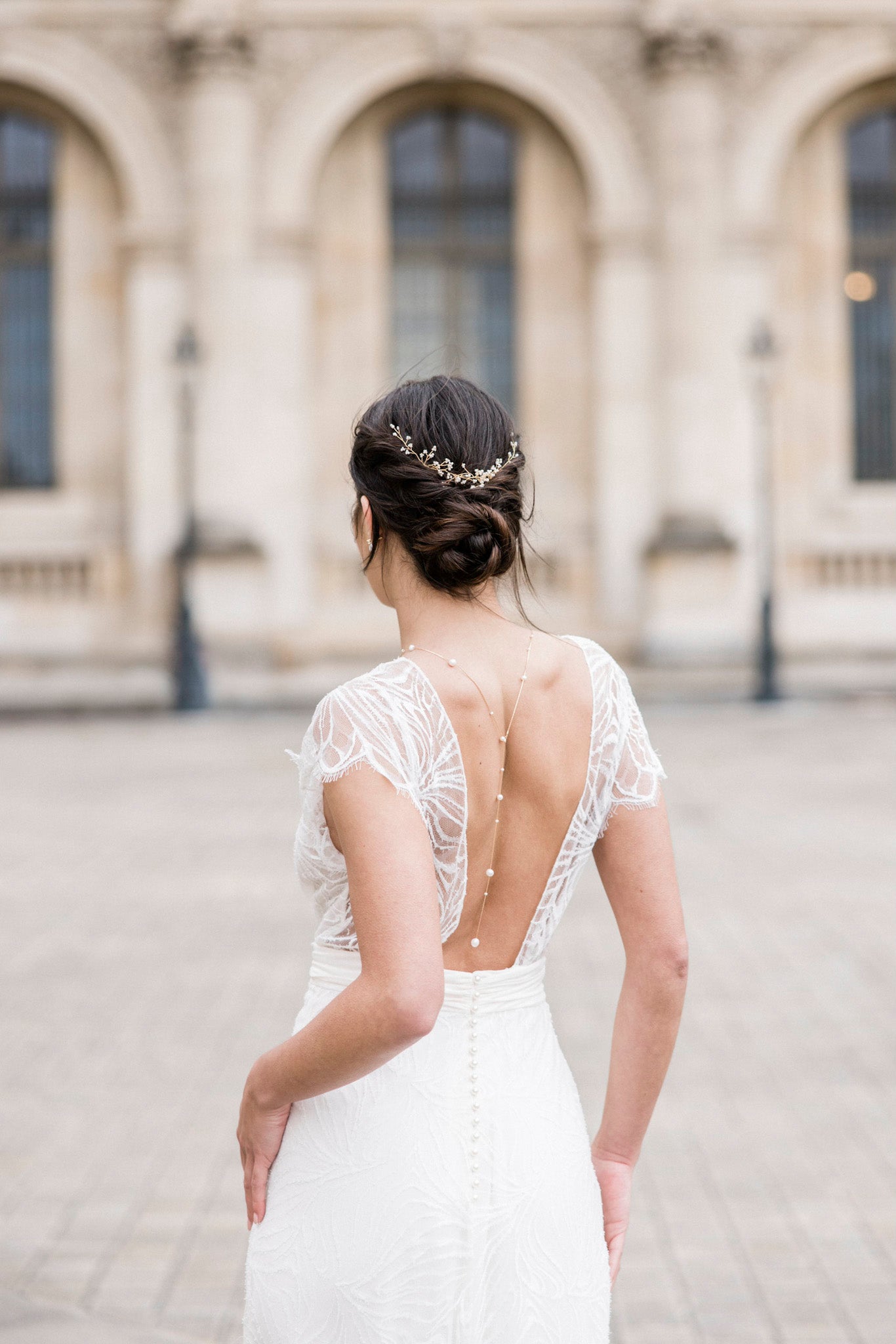 Mariée de dos sur la place du louvre à Paris portant sur son chignon bohème une vigne à cheveux doré à perles et un collier de dos à perles assorti