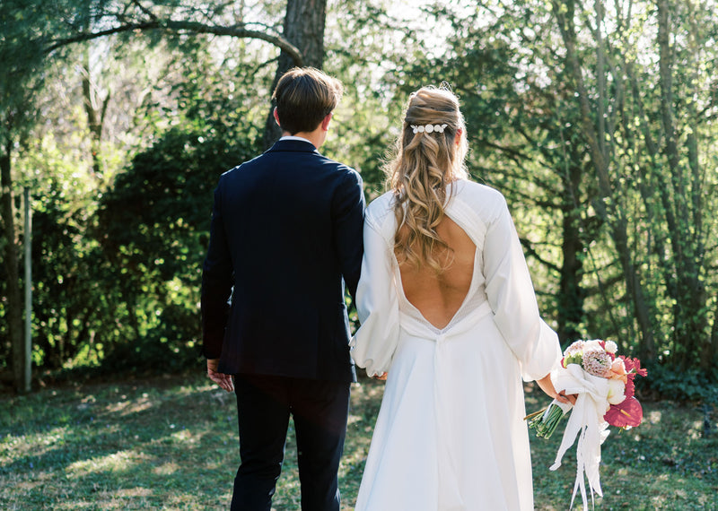 Couple de mariés de dos dans un jardin, homme avec un costume bleu foncé et femme avec une robe blanche à décolleté de dos, bouquet de fleur rose et blanc et peigne à cheveux avec des pièces rondes arrondies blanches