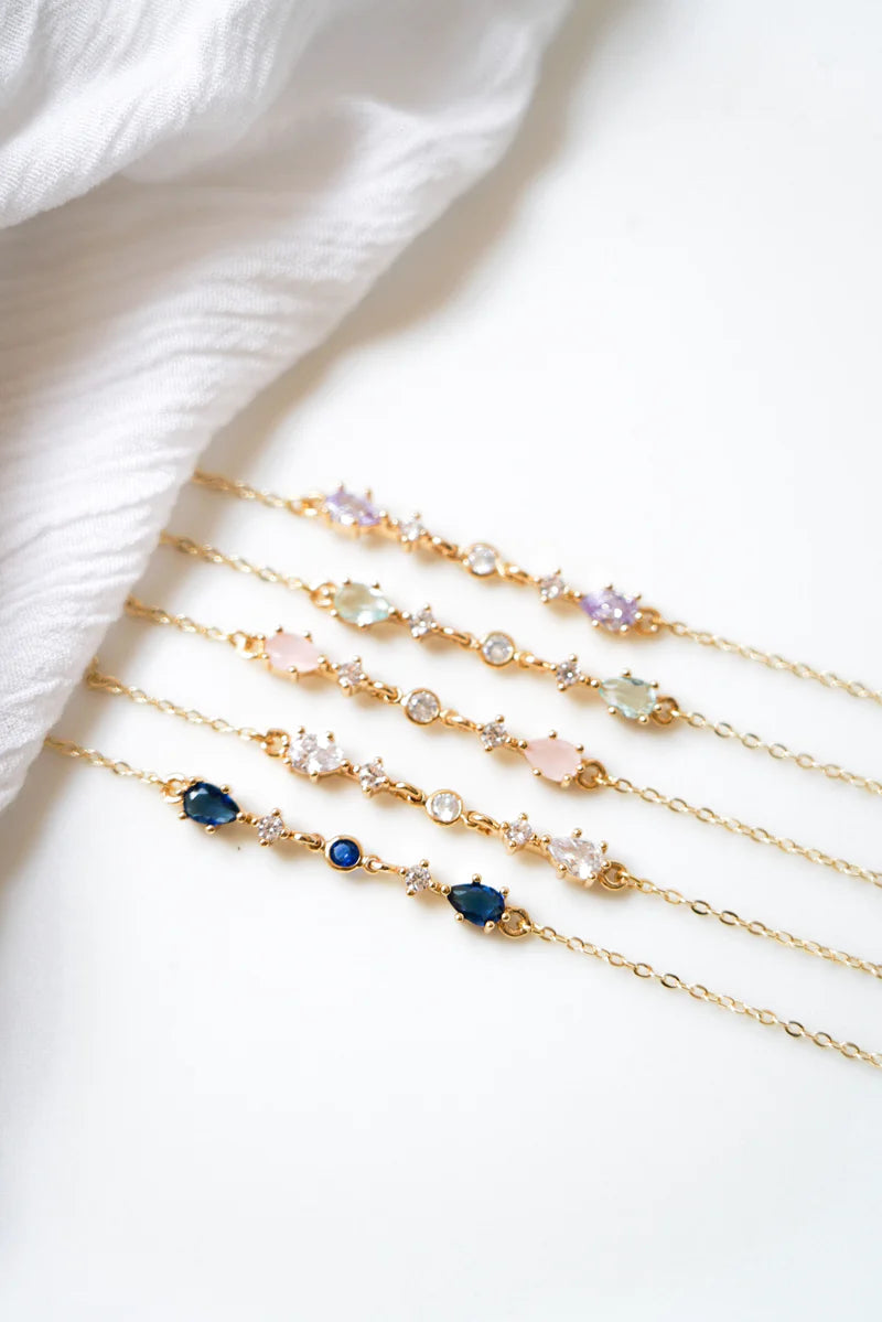 Cinq bracelet faits de chaines dorées et chacun avec deux pierres de couleurs différentes : bleues, blanches, roses, vertes et violettes