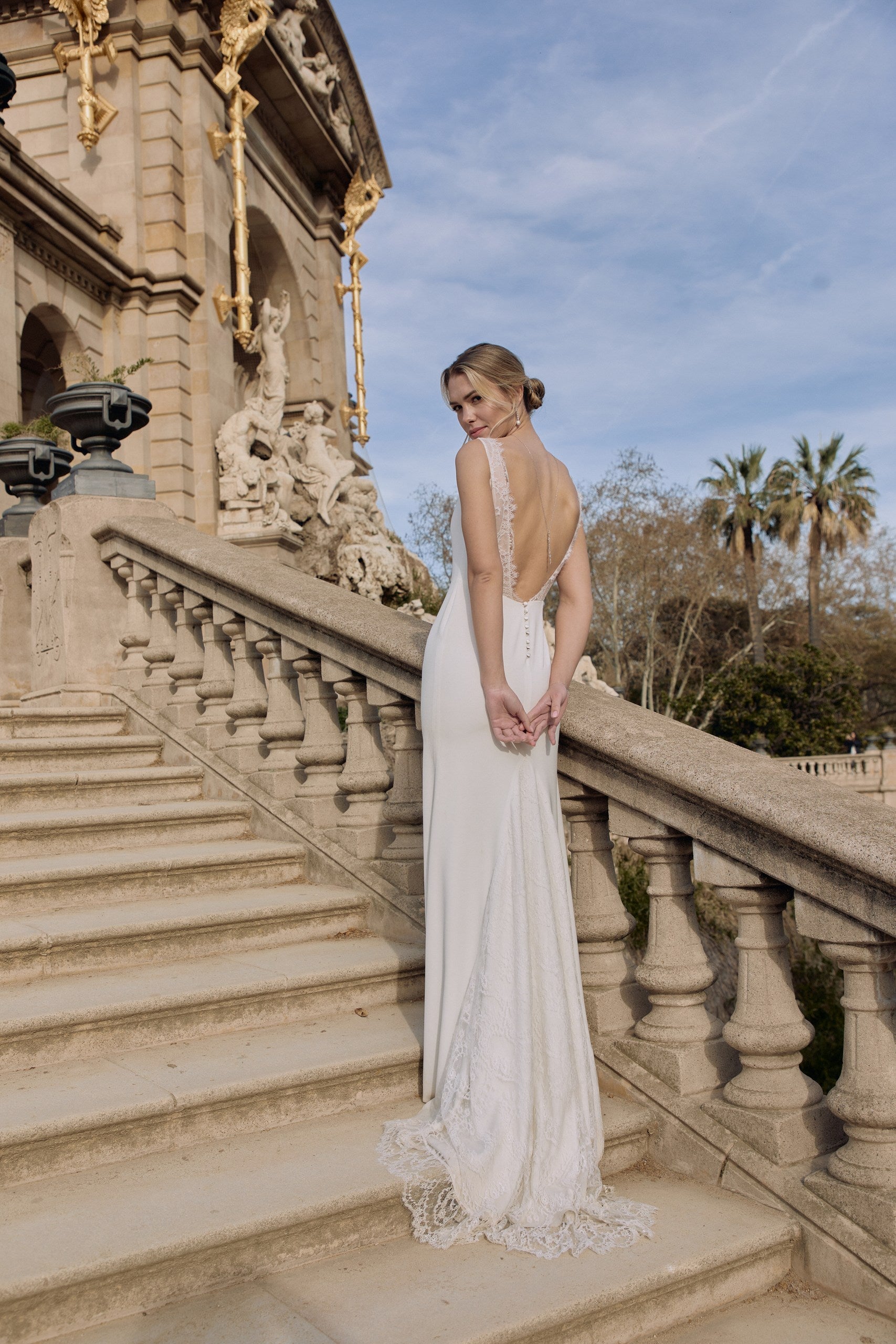 Jeune femme de loin sur des escaliers se tenant les mains de dos donnant une allure romantique à sa tenue et ses bijoux de mariage.