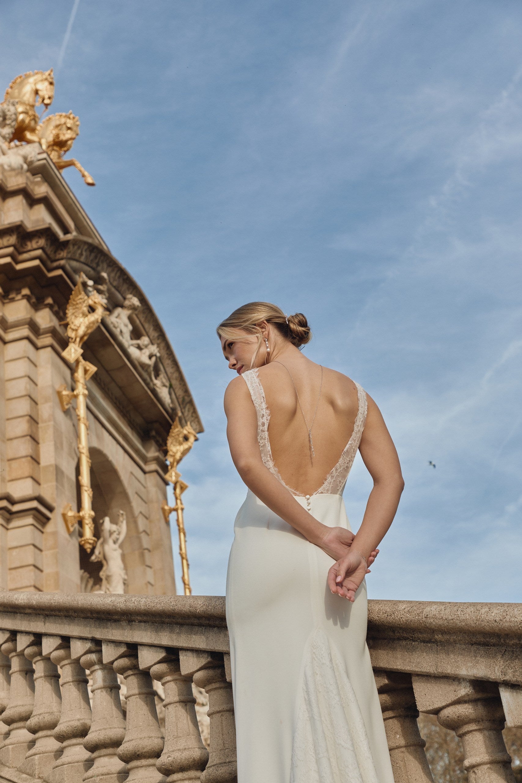 Femme de dos se tenant les mains avec une robe de mariée à grand décolleté de dos montrant une parure de bijoux raffinée