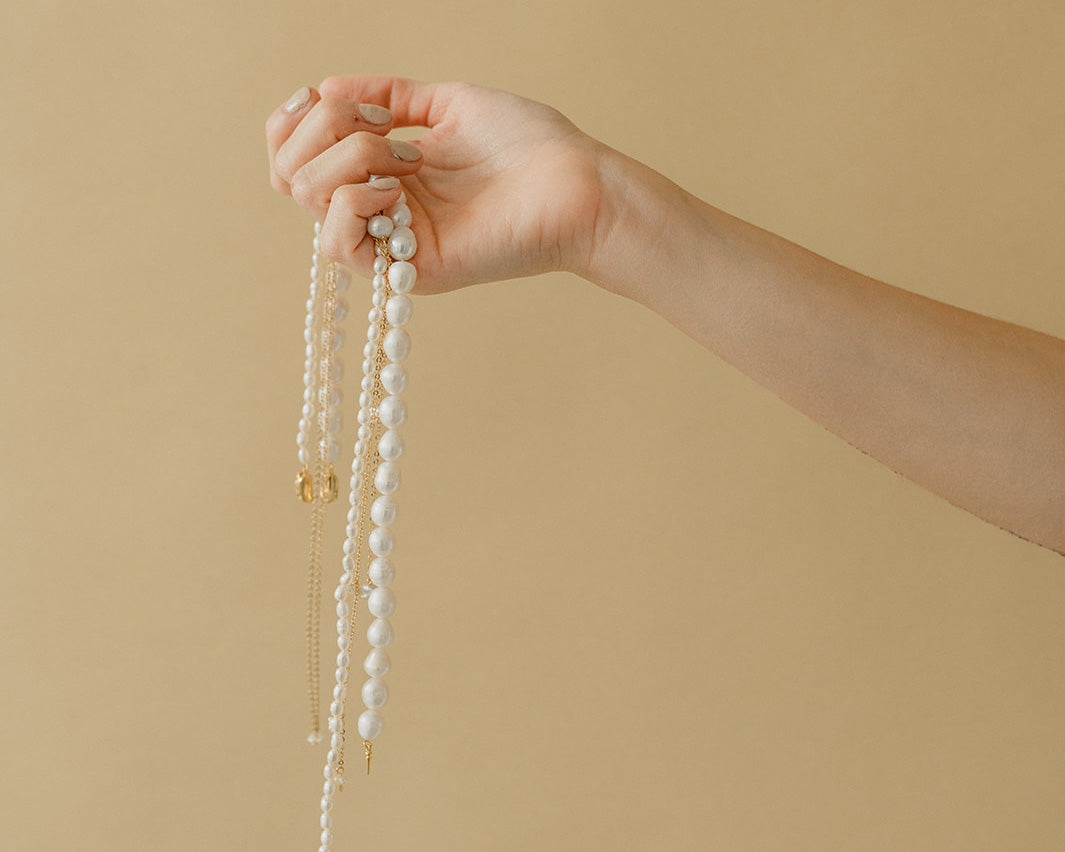 femme portant dans sa main des colliers de perles naturelles de tailles différentes pour mariées sur un fond beige