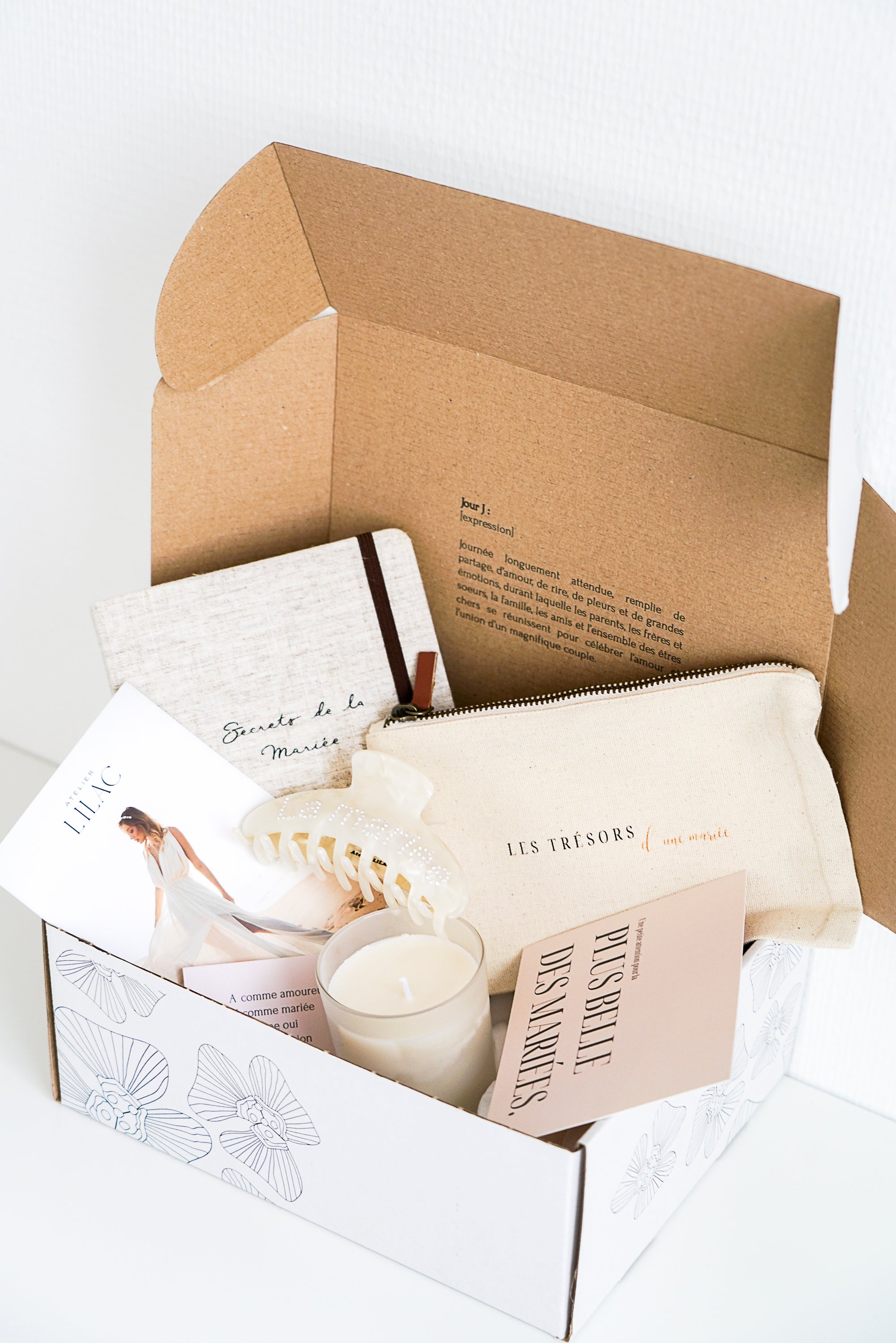 Box en carton blanc imprimé de fleurs remplis de cadeaux de mariage, bougie, carnet, trousse, peigne, cartes, guide de mariage