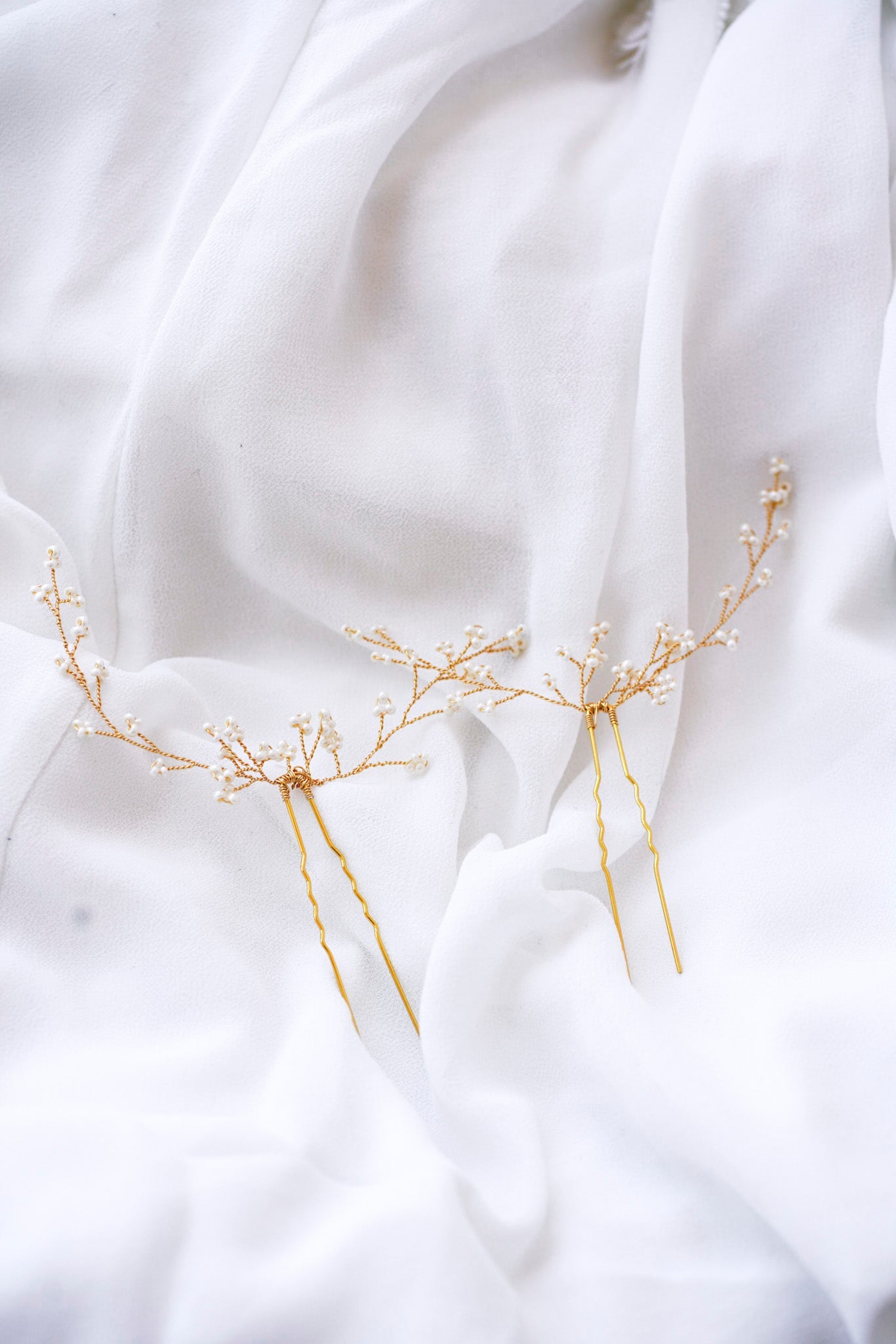 Deux pics à cheveux dorées formant des branches fleuries de petites fleurs en perles rocailles, posés sur un drap blanc