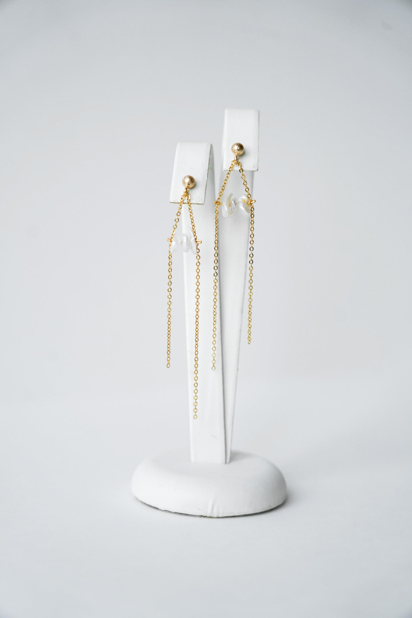 une paire de boucles d'oreilles pour une mariée composé de petites perles naturelles et de chaines en or sur un portant blanc à bijoux 