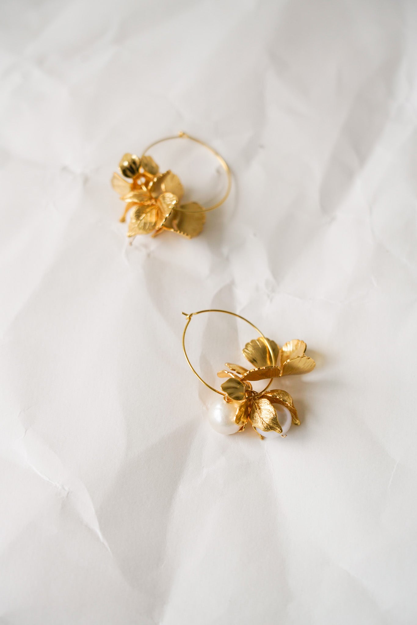 Créoles fines et élégantes posées sur fond blanc, ornées d’une petite fleur mobile suspendue sur chaque anneau, accompagnée d’une perle de culture blanche délicatement attachée. Un bijou raffiné et léger, alliant mouvement et douceur pour une allure romantique.