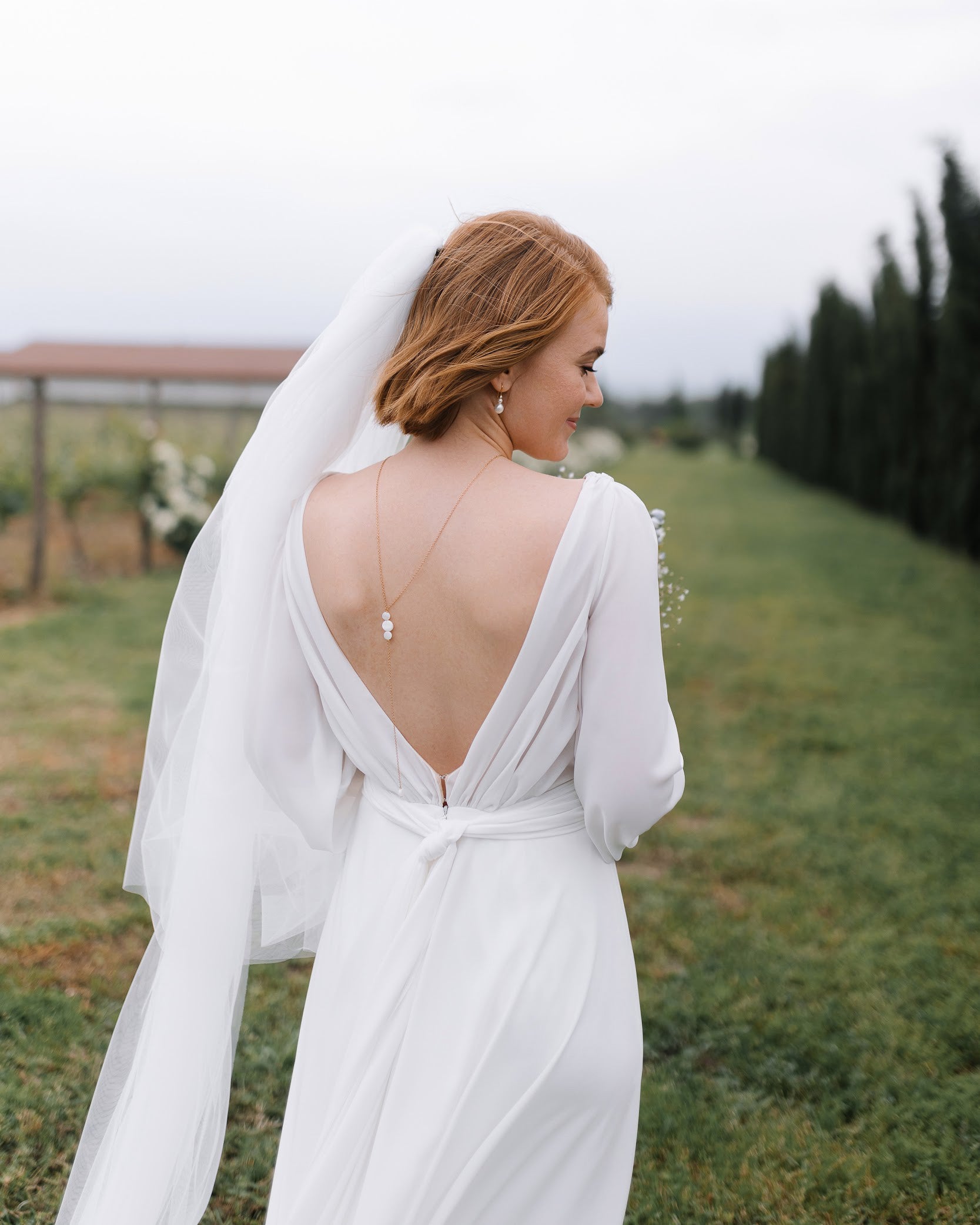 mariée en robe blanche à voile avec un décolleté de dos portant un collier de dos en nacre et son bouquet dans les mains