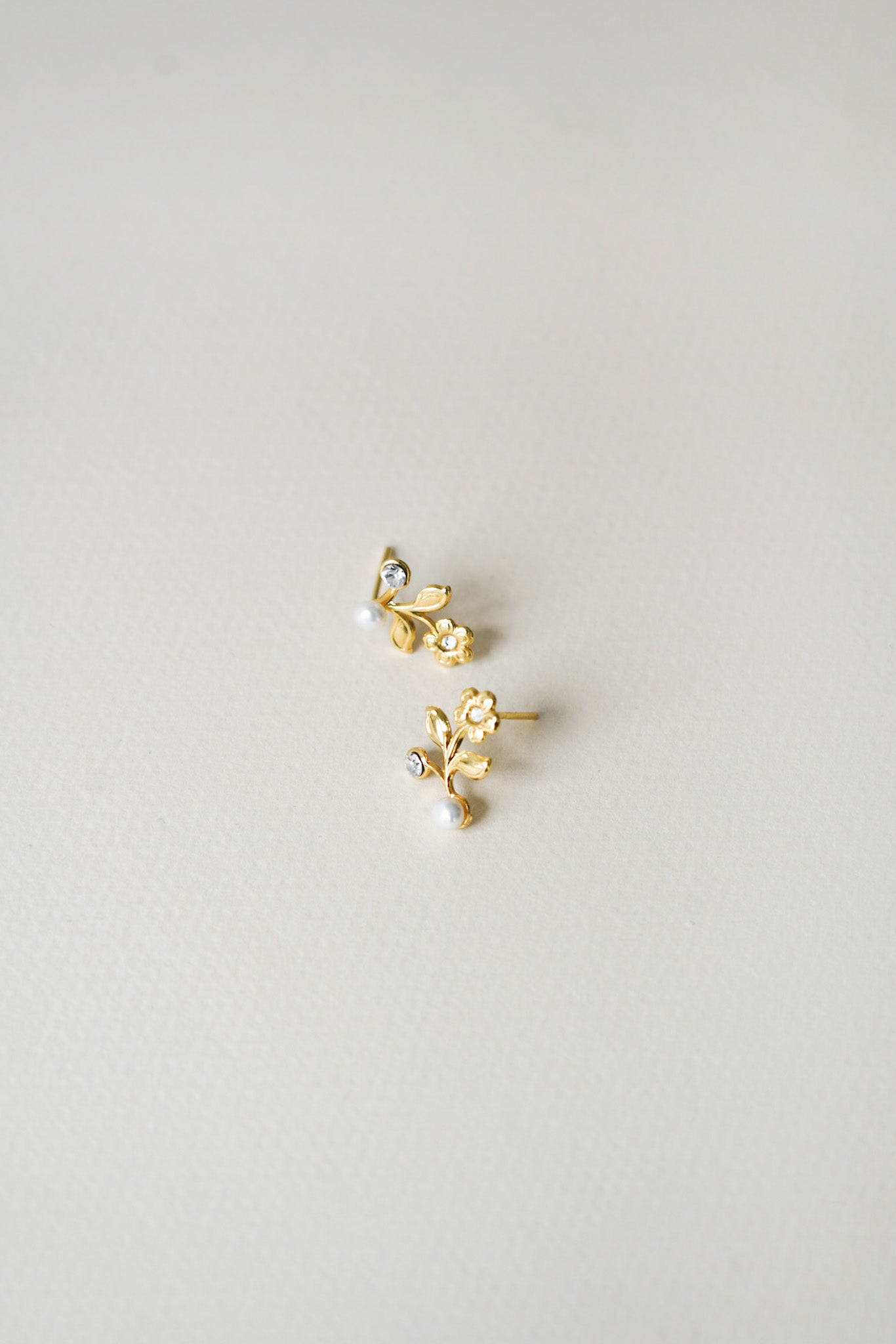 Petites boucles d’oreilles délicates en forme de fleur, ornées en leur centre d’un cristal étincelant couleur argent. Une perle élégante est délicatement posée à la base de chaque boucle, créant un contraste raffiné sur un fond blanc doux vu de côté. Le design floral apporte une touche romantique et lumineuse, parfait pour un mariage ou une occasion spéciale.