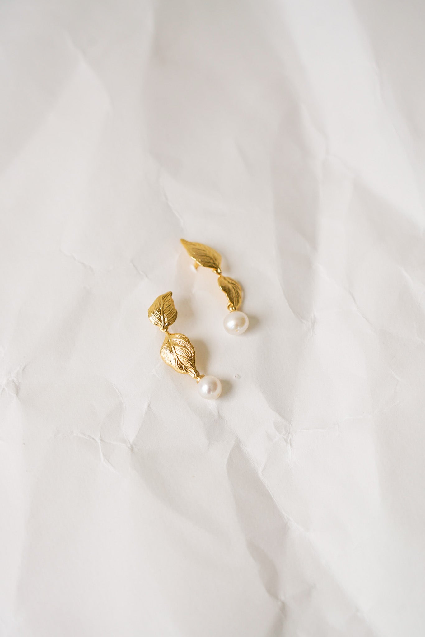 Boucles d’oreilles délicates posées sur un papier froissé, composées de deux petites feuilles en forme de lune, l’une orientée vers le haut et l’autre vers le bas, avec une perle blanche suspendue entre elles. Un design fin et élégant, parfait pour apporter une touche naturelle et raffinée à une tenue. 