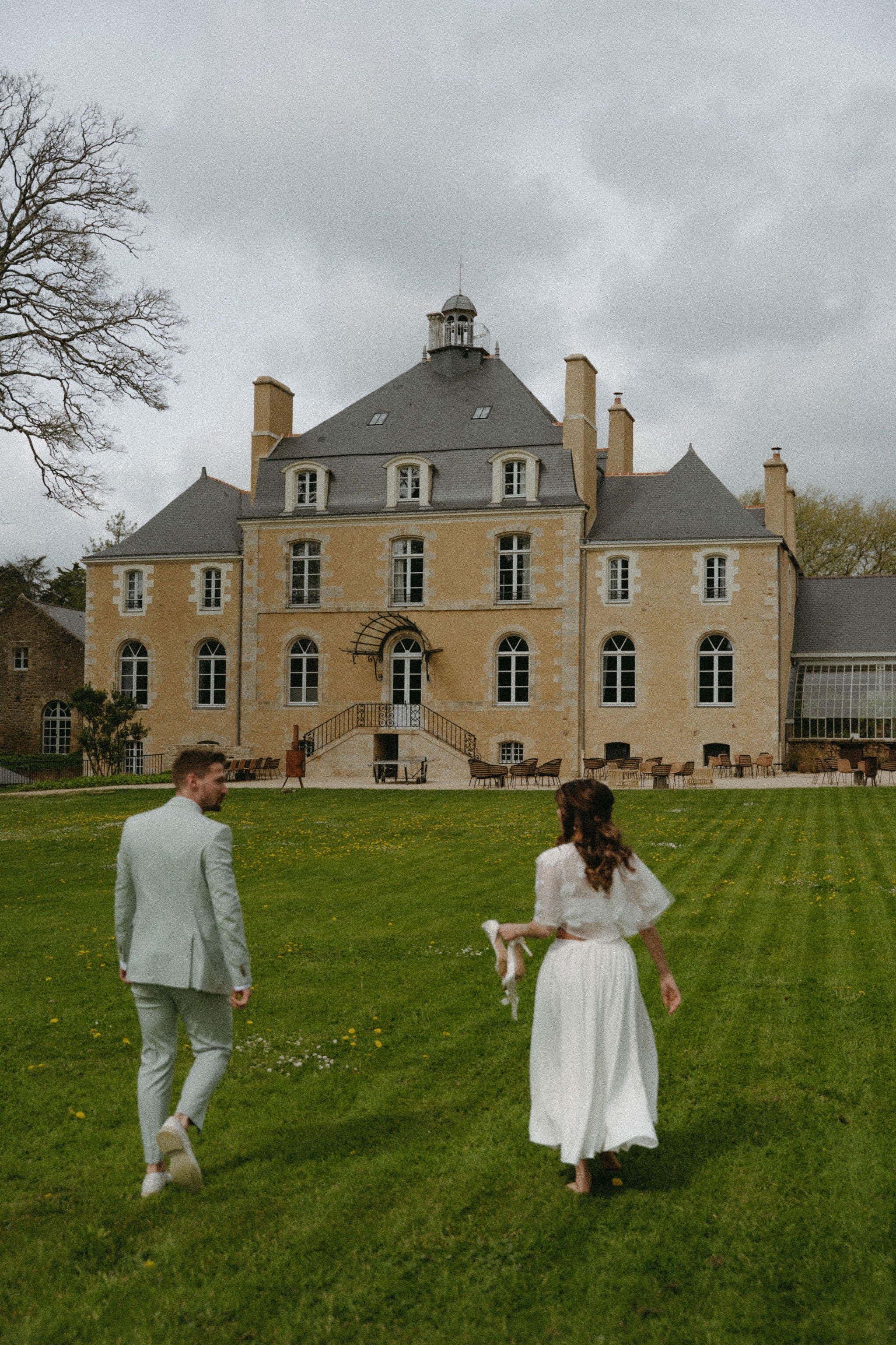 Photo de couple marié marchant sur la pelouse devant château, robe mariée élégante, mariage château France, marié en costume, photo romantique