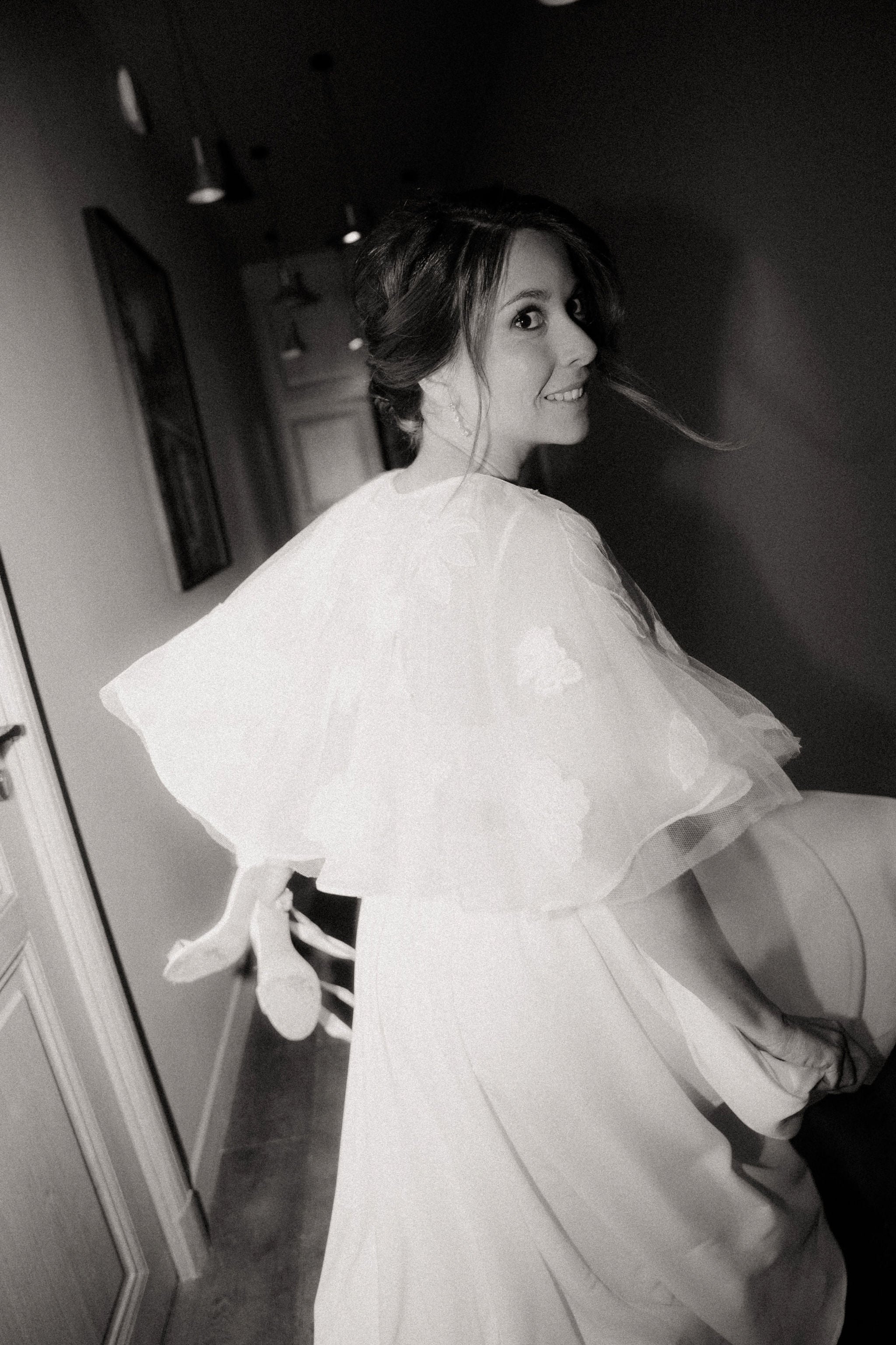Photo en noir et blanc de mariée qui court dans un couloir en tenant ses chaussures à talons dans une main, mariée qui sourit, photo prise sur le vif de mariée heureuse. 