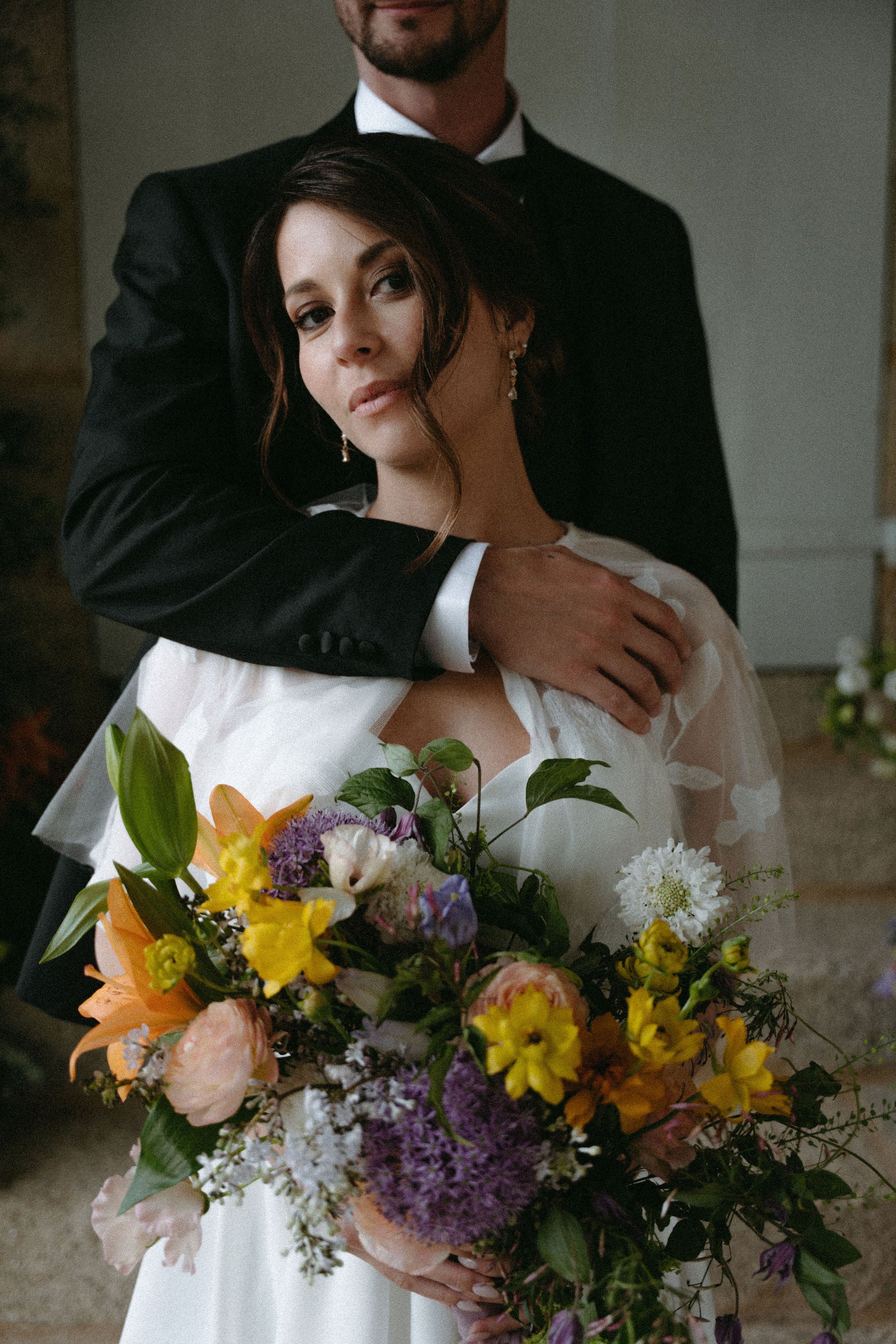 Photo romantique d'un couple marié, homme en costume qui enlace sa femme en robe de mariée, mariée élégante qui tient un bouquet de fleurs et qui porte les boucles d'oreilles vitoria Atelier Lilac, arrangement floral mariage, fleurs mariage, bouquet mariage, mariage élégant en château 