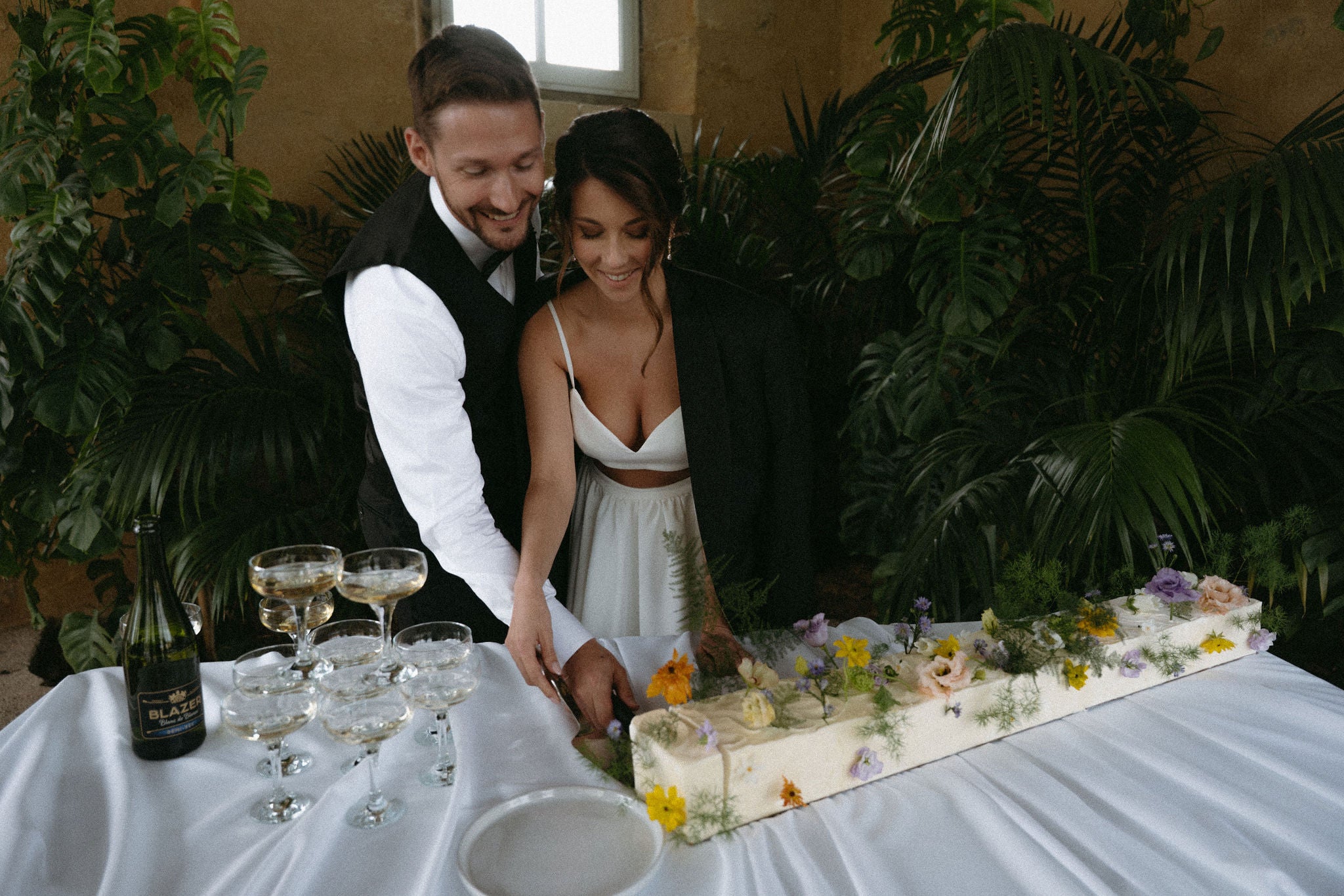 Couple marié souriant qui coupe une part de gâteau de mariage avec du champagne, mariage élégant, wedding cake, gâteau mariage, couple, décor végétal, réception au château, photo romantique mariage élégant 