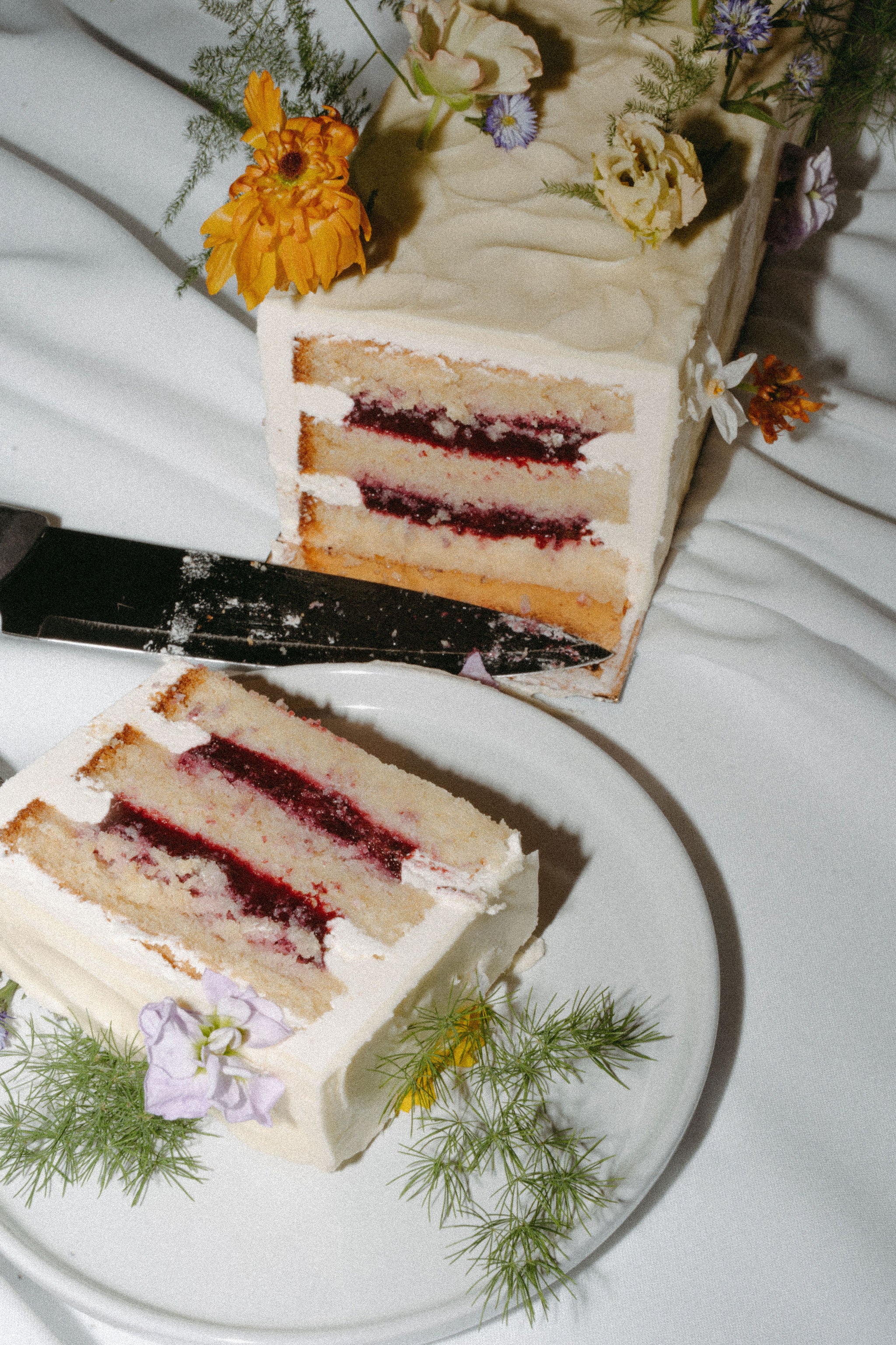 Photo esthétique éditoriale de gâteau de mariage coupé dans une assiette sur une table avec des fleurs et un couteau, le gâteau est  la crème et aux fruits rouges, gâteau de mariage, wedding cake, gâteau minimaliste, cake design, mariage élégant en château 
