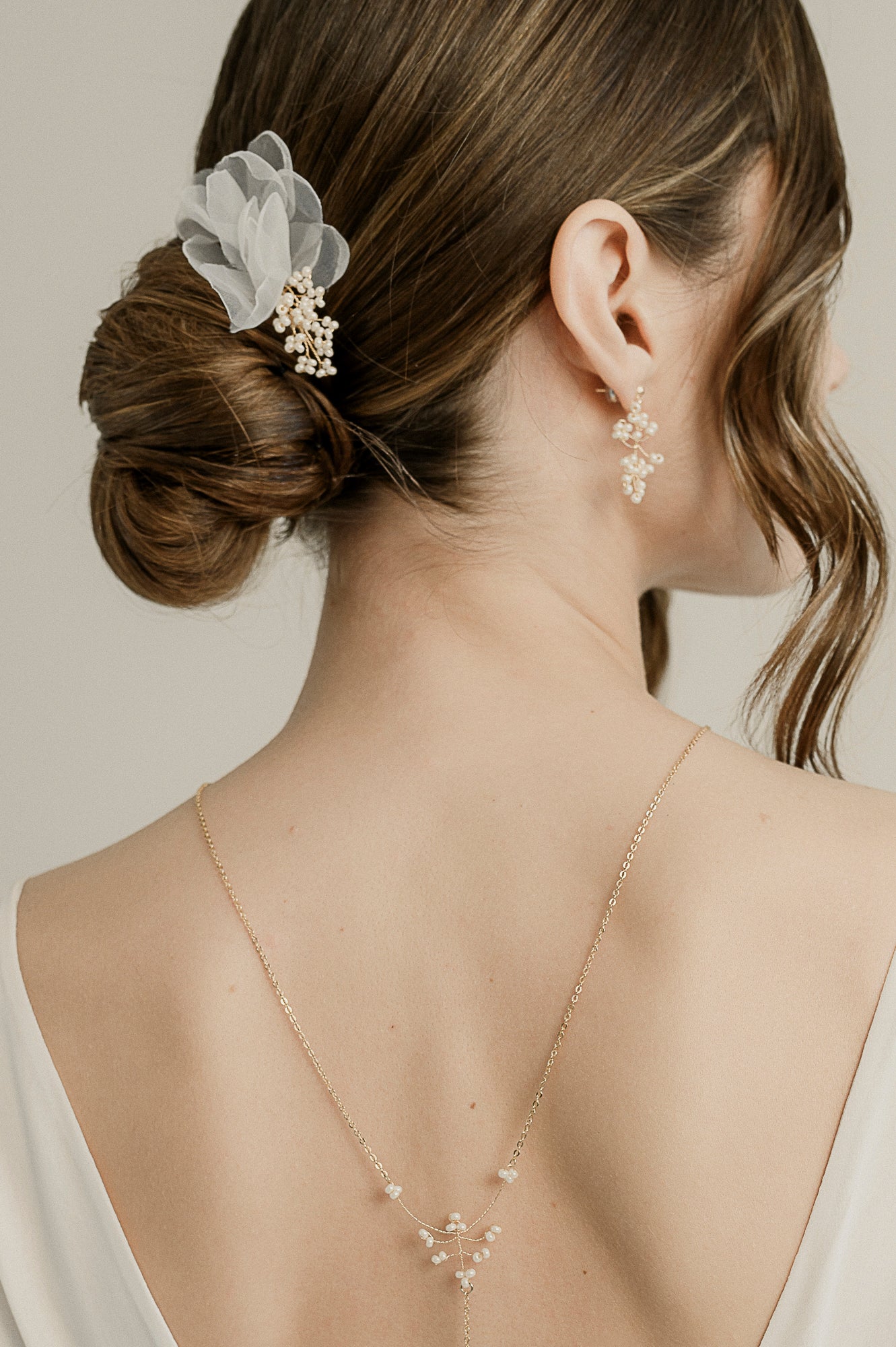 Femme de dos avec un chignon de mariée montrant des accessoires de cheveux, un collier de dos et des boucles d'oreilles en perles rocailles