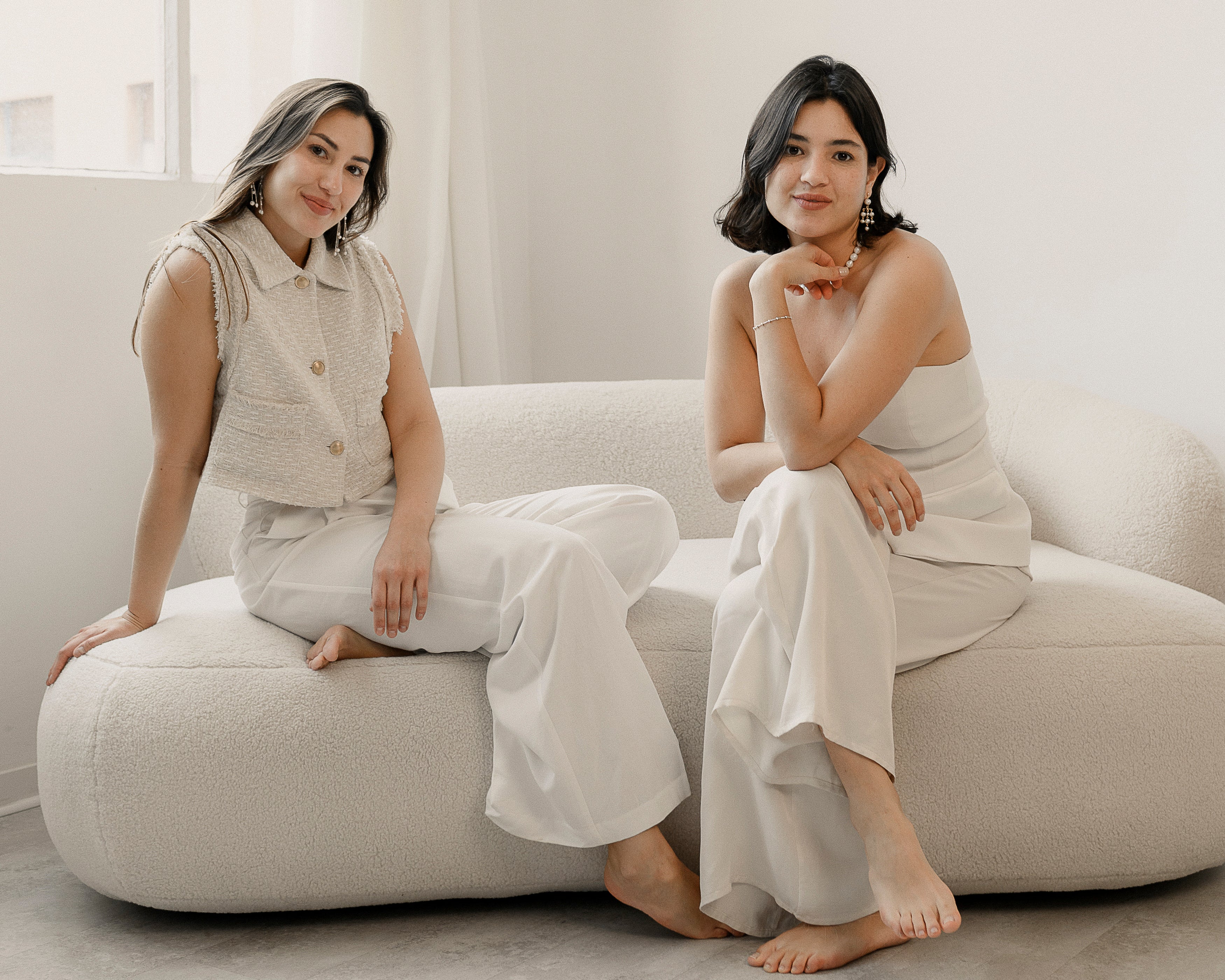 Les deux fondatrices d'Atelier Lilac assises sur canapé portant du blanc et touchant leurs collier et autres bijoux de mariées