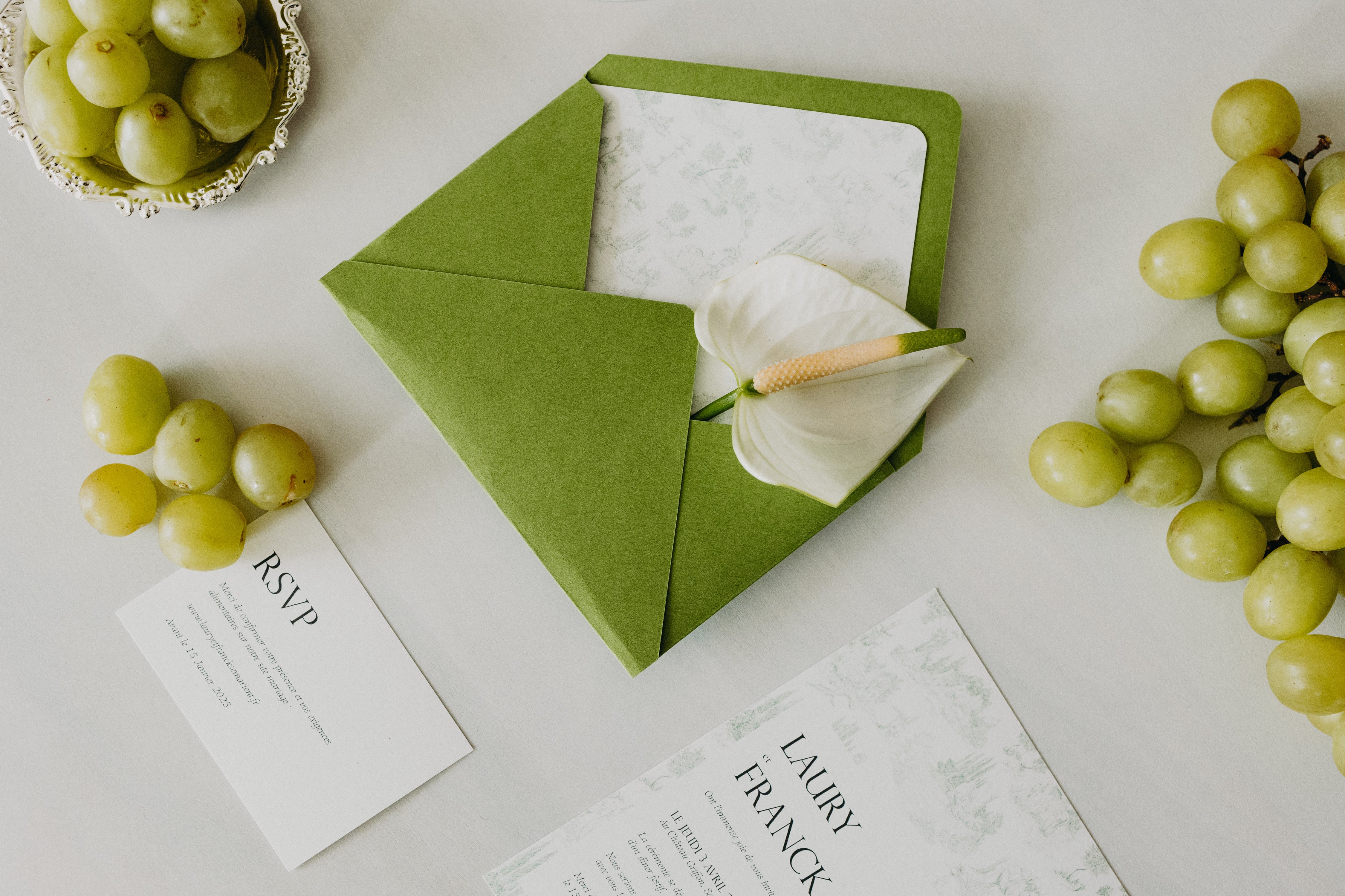 Papèterie sur le thème matcha et olive, enveloppe couleur matcha accompagnée d'élégants et modernes RSVP et faire parts ainsi que d'une fleur et de raisins.