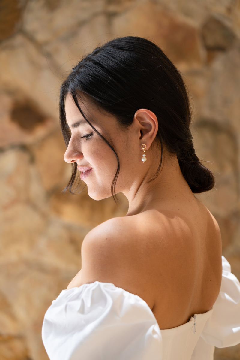 Mariée souriante avec robe de mariée minimaliste aux épaules dénudées et boucles d'oreilles à double anneaux entrelacés et deux perles naturelles blanches