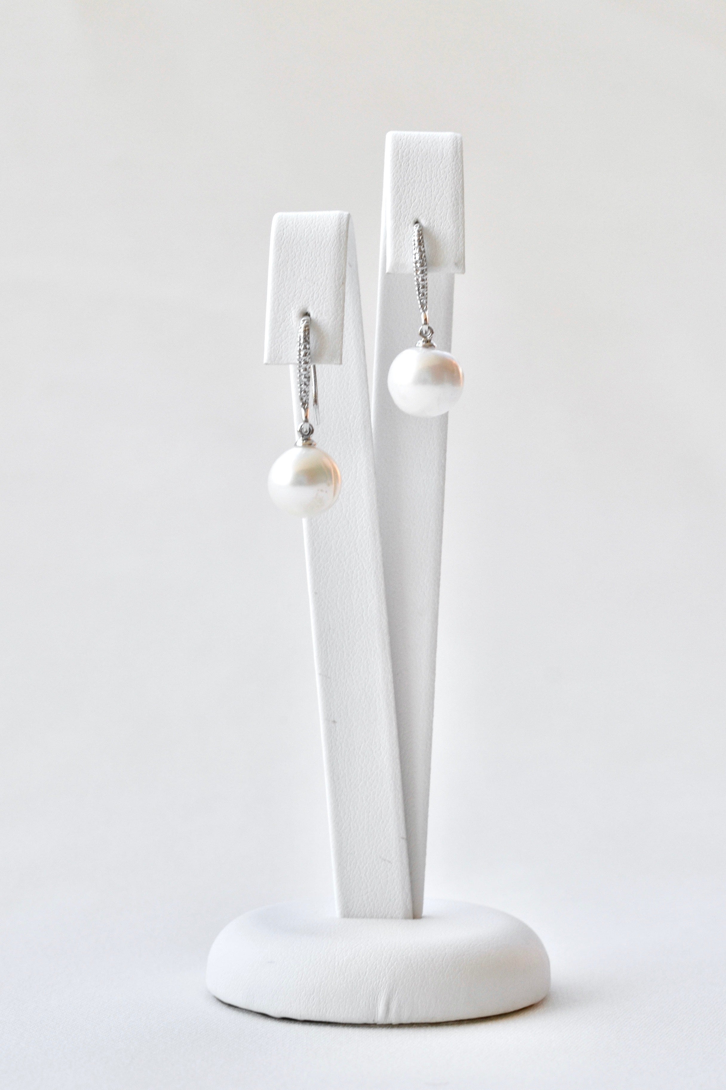 boucles d'oreilles avec une attache en argent et des petites cristaux, une perle pendant de cette attache et les boucles sont sur un socle à bijoux blanc