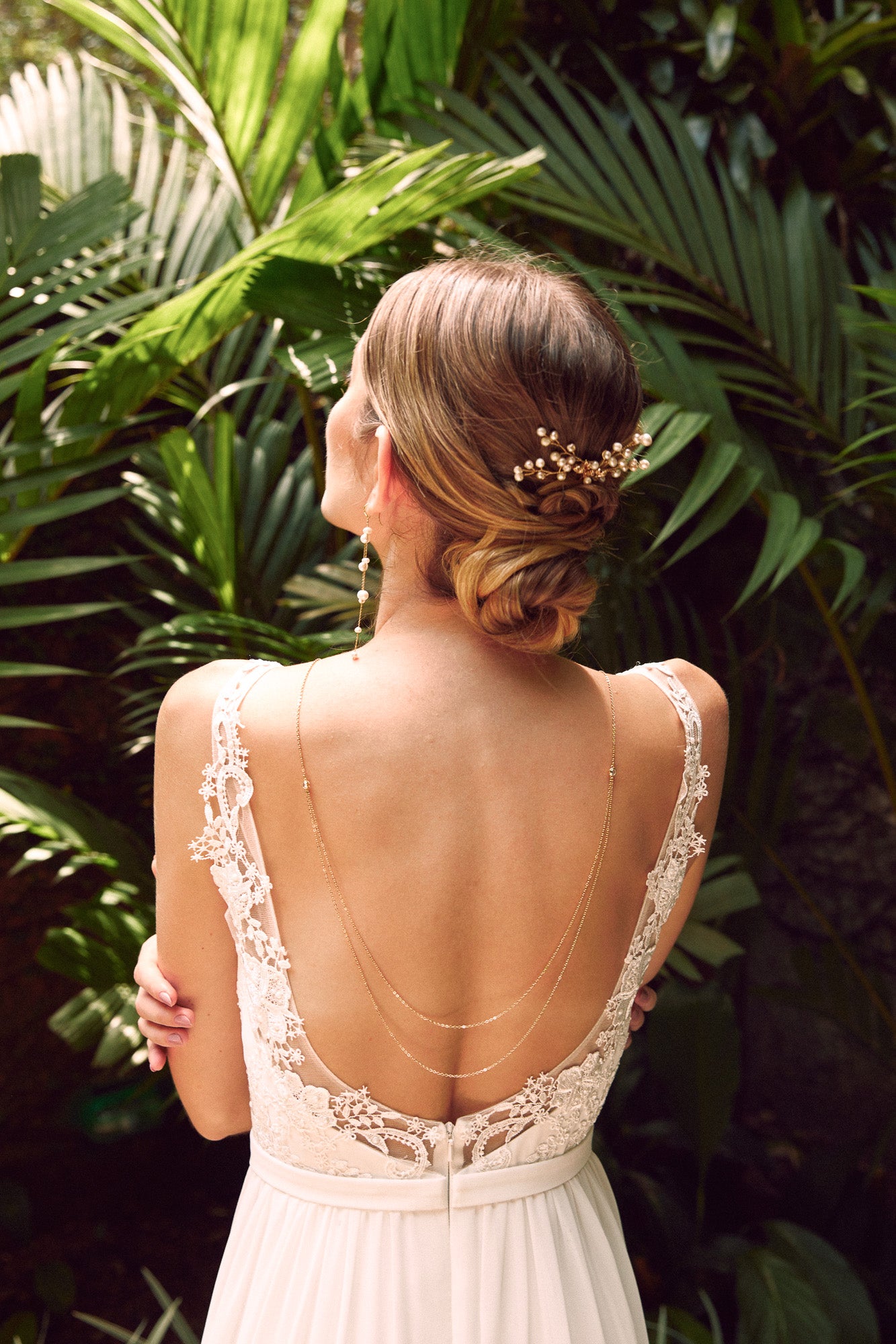 Mariée de dos devant un jardin tropical portant un collier de dos à doubles chaines en or, des boucles d'oreilles pendantes en perles et un pic à cheveux bohème en perles naturelles